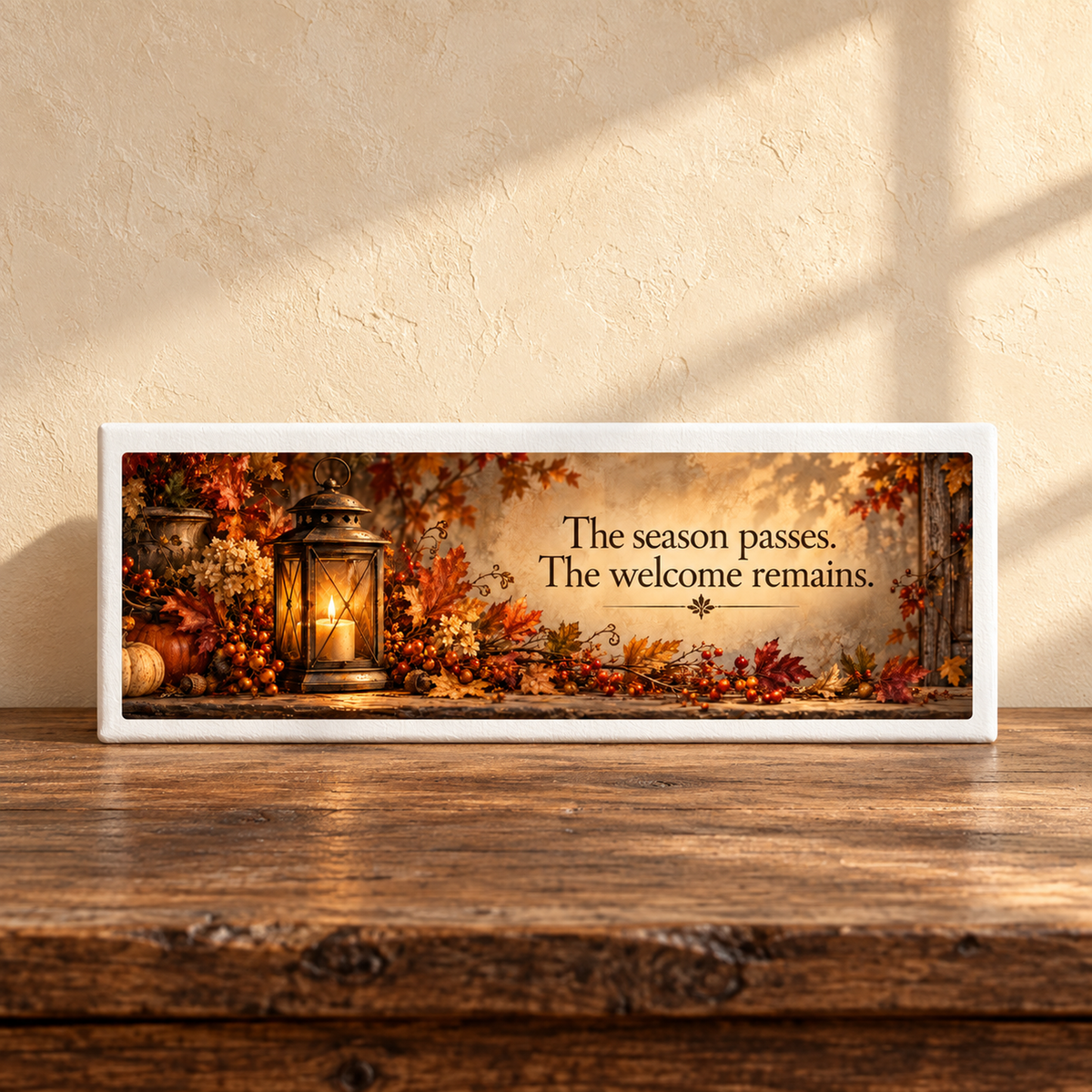 The Welcome Remains Autumn Shelf Sitter by Mirabilia Boutique sits on a wooden table with autumn leaves and a lantern, sunlight casting warm shadows behind. Its text reads: “The season passes. The welcome remains.”.