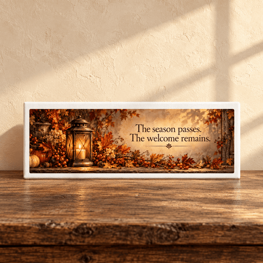 The Welcome Remains Autumn Shelf Sitter by Mirabilia Boutique sits on a wooden table with autumn leaves and a lantern, sunlight casting warm shadows behind. Its text reads: “The season passes. The welcome remains.”.