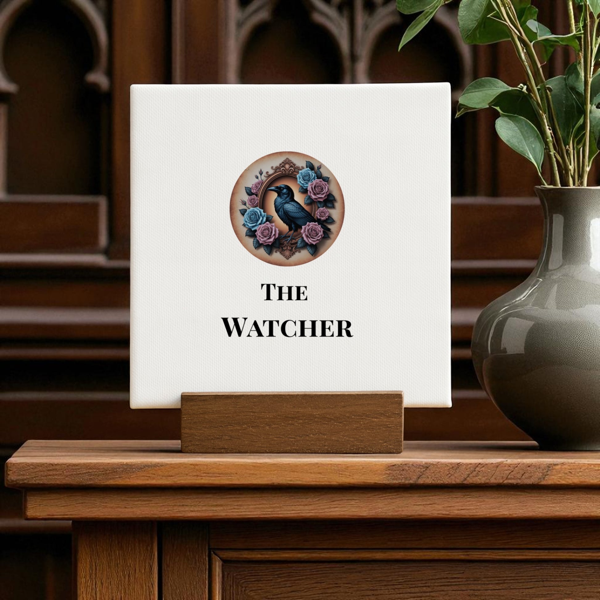 The Mini Gothic Sign | The Watcher by Mirabilia Boutique features raven and rose art on a white square sign with a wooden stand. A gray vase with green leaves complements the dark wood gothic décor.