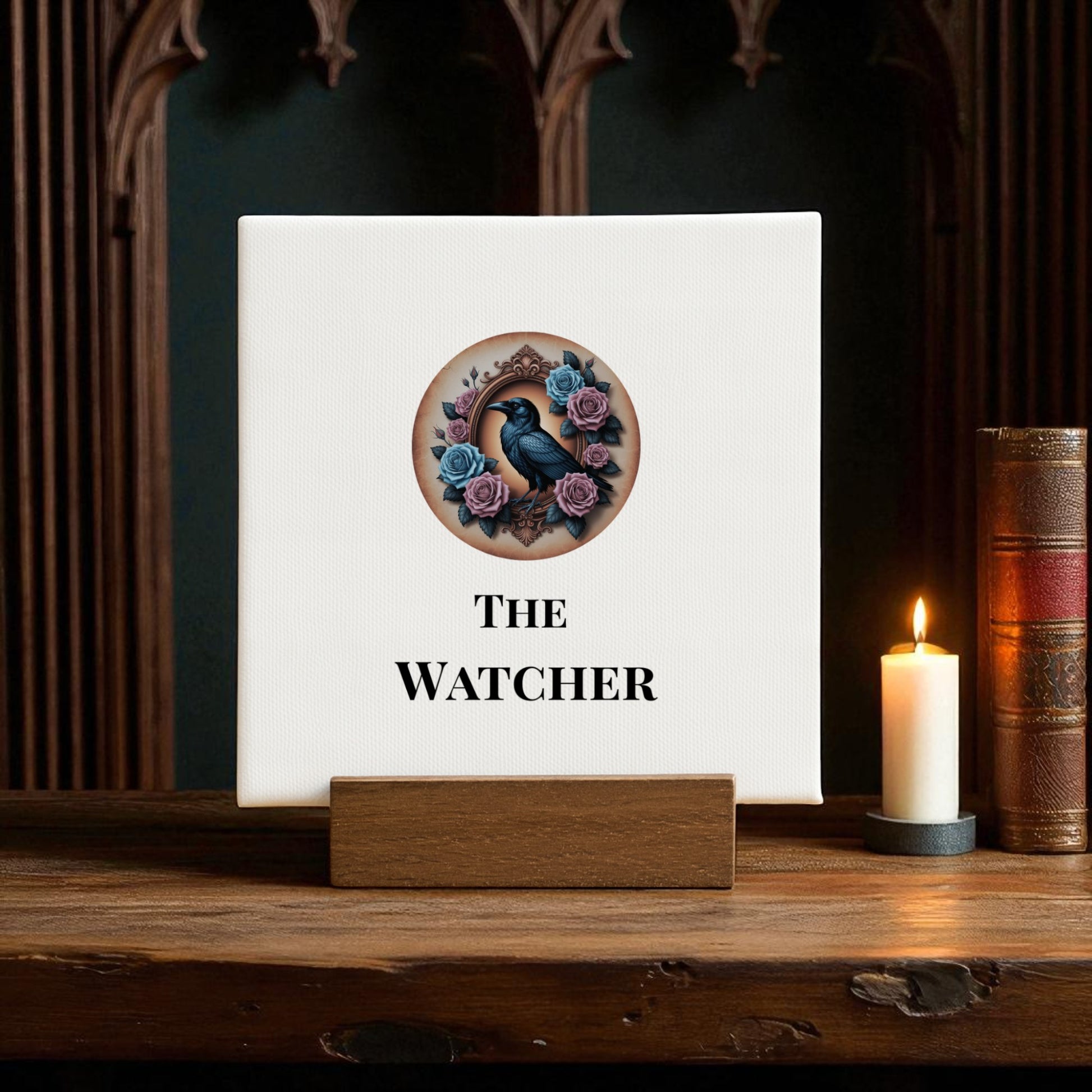 The Mini Gothic Sign by Mirabilia Boutique, titled "The Watcher," features a black raven with roses on a square canvas stand—evoking dark romanticism and raven lore—beside a lit candle and antique books.