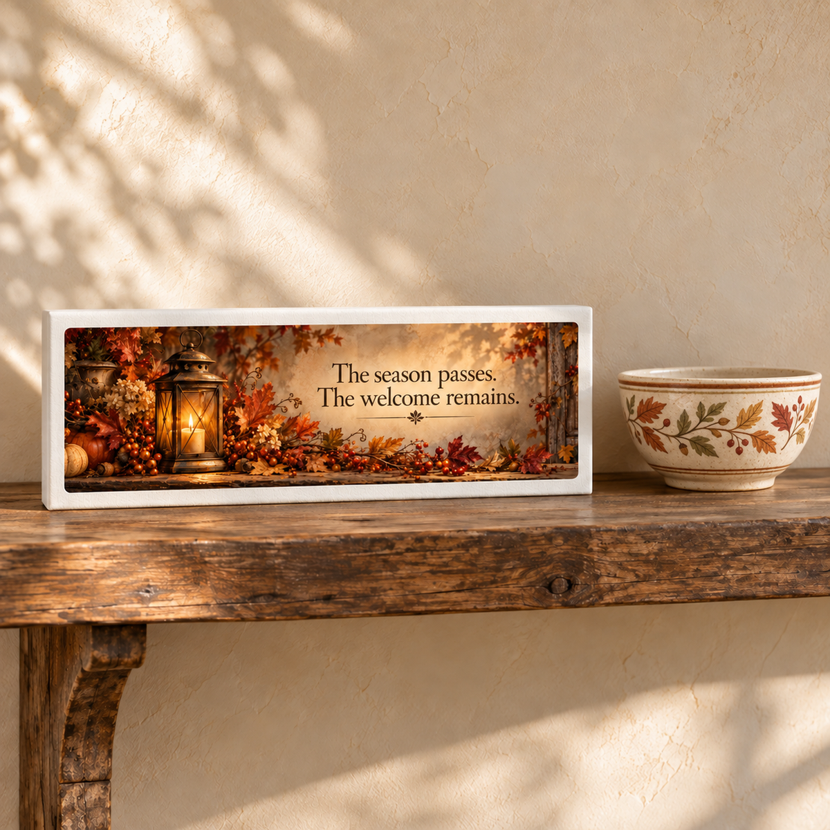 A wooden shelf displays a bowl with autumn leaf designs and Mirabilia Boutique’s The Welcome Remains Autumn Shelf Sitter—featuring a lantern, pumpkins, fall foliage, and the phrase “The season passes. The welcome remains.”.