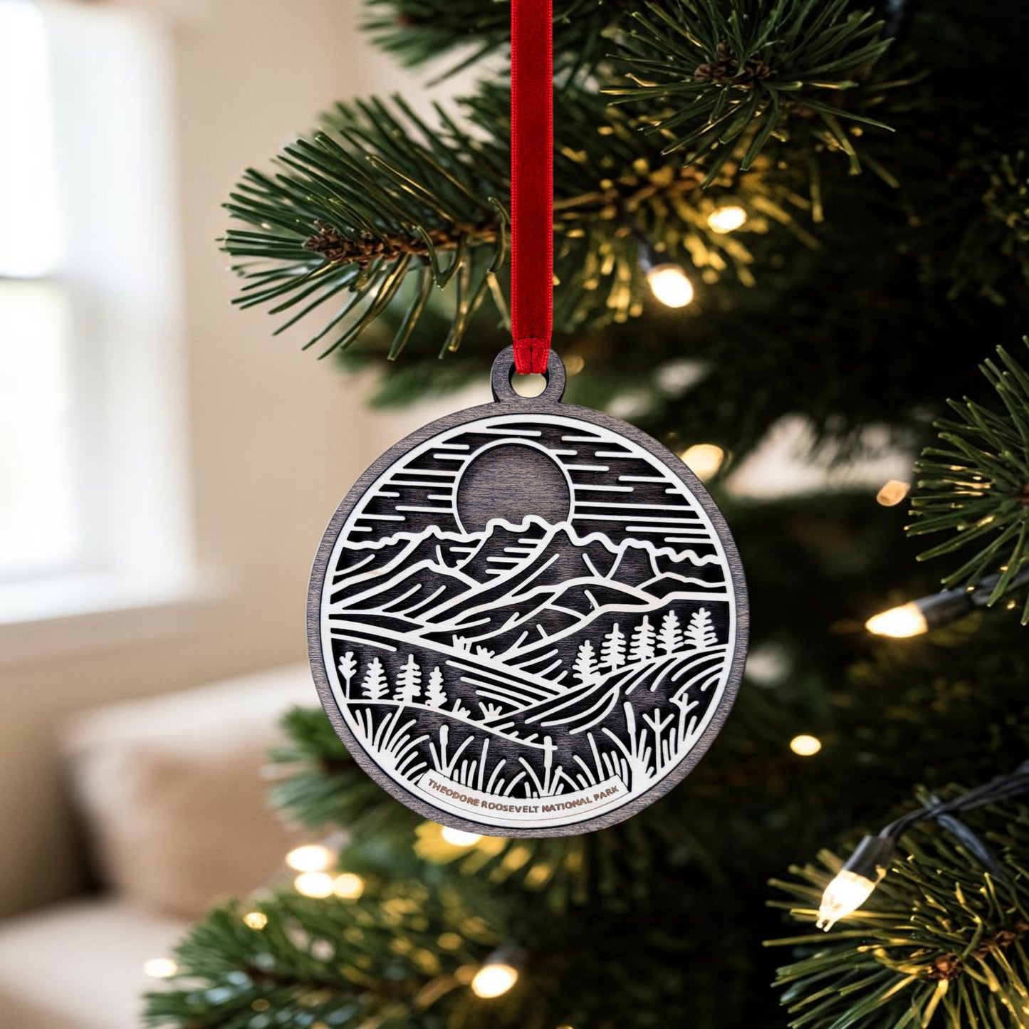 A beautiful Theodore Roosevelt National Park Line Art Christmas Ornament from Mirabilia Boutique, featuring a mountain and pine tree scene in silver, hangs from a red ribbon on a Christmas tree adorned with white lights—an eye-catching choice for any nature lover.