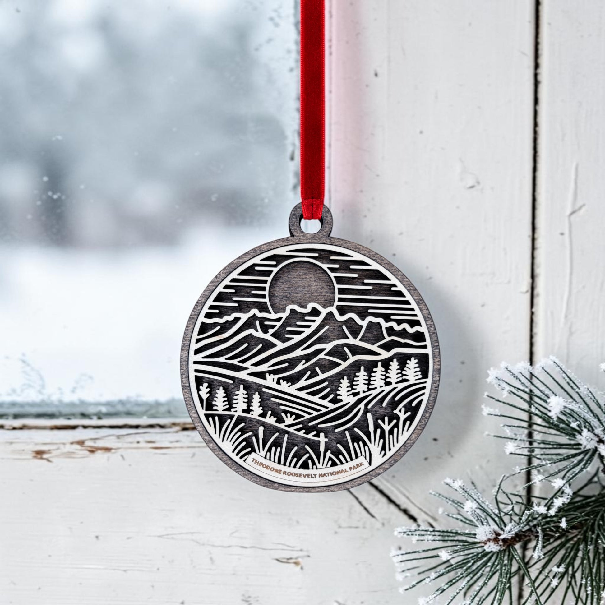A round metal ornament from Mirabilia Boutique, featuring line art of mountains, trees, and a moon inspired by Theodore Roosevelt National Park, hangs by a red ribbon on a snowy window beside pine branches—perfectly capturing the spirit of the Theodore Roosevelt National Park Line Art Christmas Ornament.