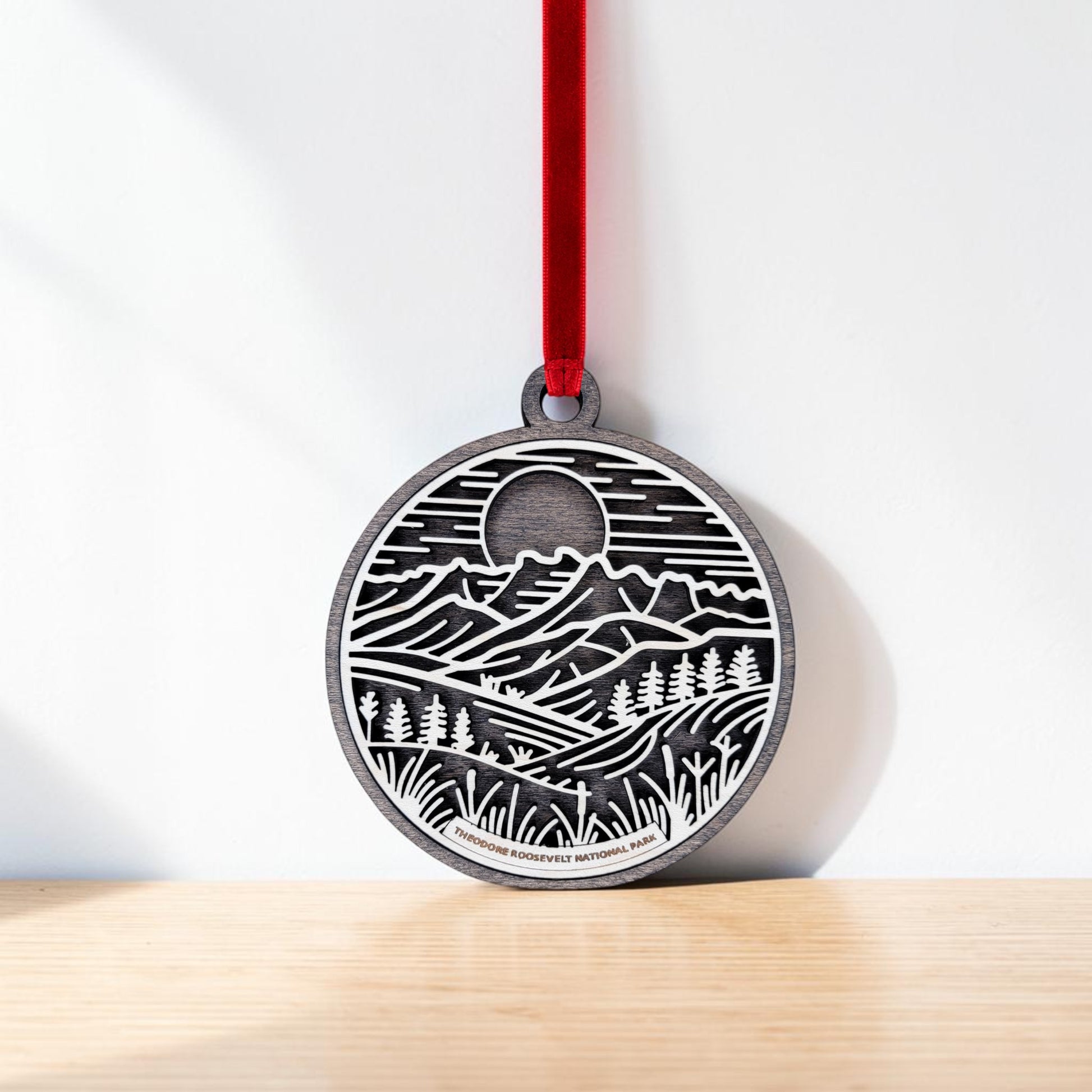 The Theodore Roosevelt National Park Line Art Christmas Ornament by Mirabilia Boutique is a round ornament with a red ribbon, featuring a black and white landscape of mountains, trees, and a sun or moon. Resting on a light wooden surface against a white wall, this wooden line art ornament makes a striking badlands keepsake inspired by the beauty of Theodore Roosevelt National Park.