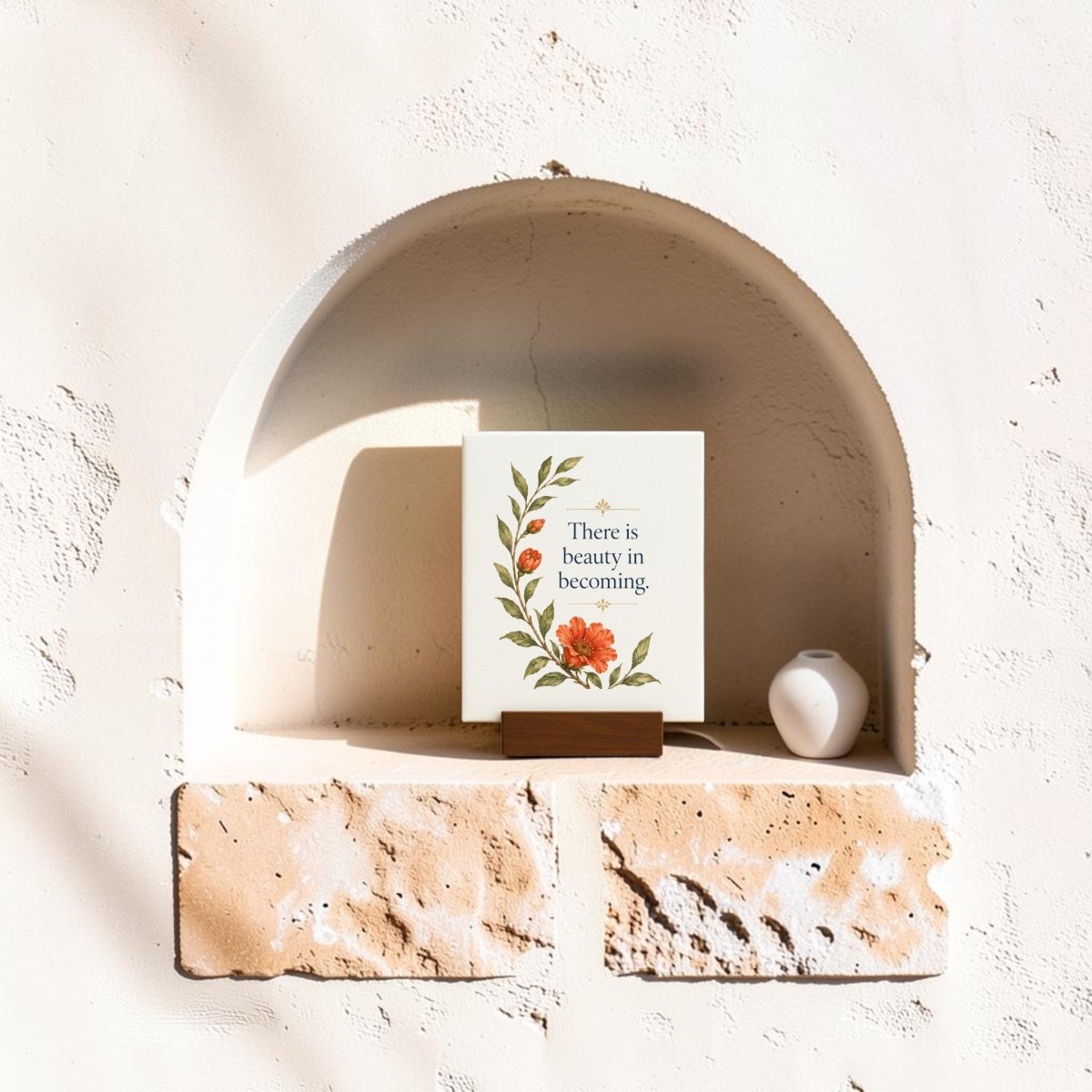 A Mirabilia Boutique “There Is Beauty in Becoming” Mini Canvas Sign, featuring a floral design and inspirational message, sits in a sunlit wall alcove next to a small white vase, adding an uplifting touch to the peaceful setting.