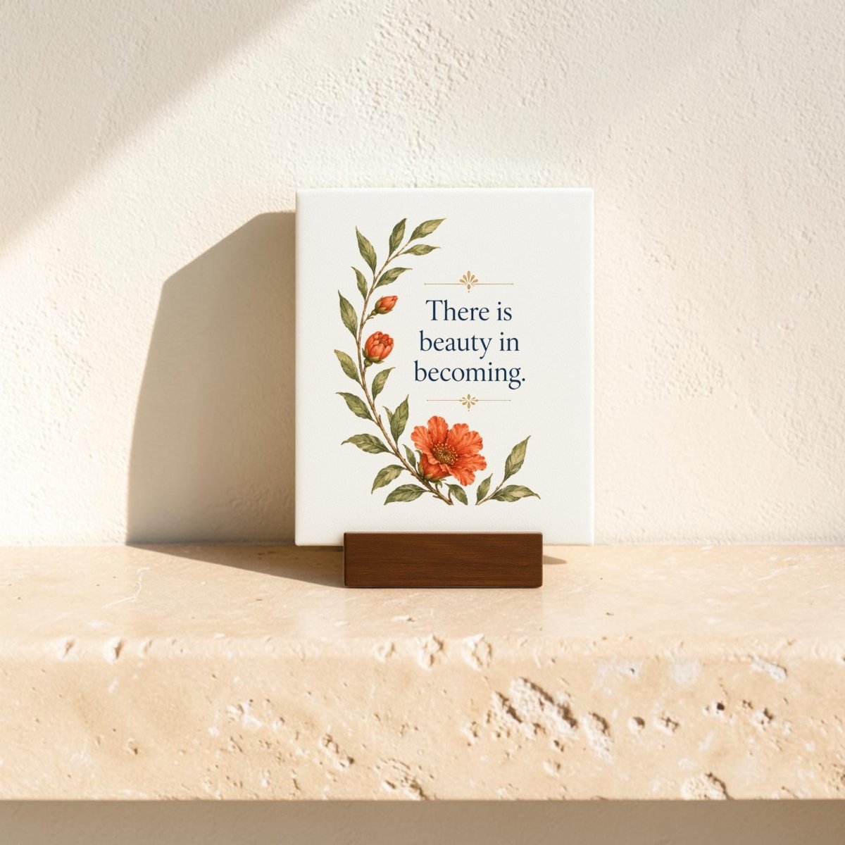 A mini canvas sign from Mirabilia Boutique, titled “There Is Beauty in Becoming,” features vibrant orange flowers and green leaves. Displayed on a wooden holder atop a beige stone surface, this botanical artwork is gently illuminated by soft sunlight in the background.