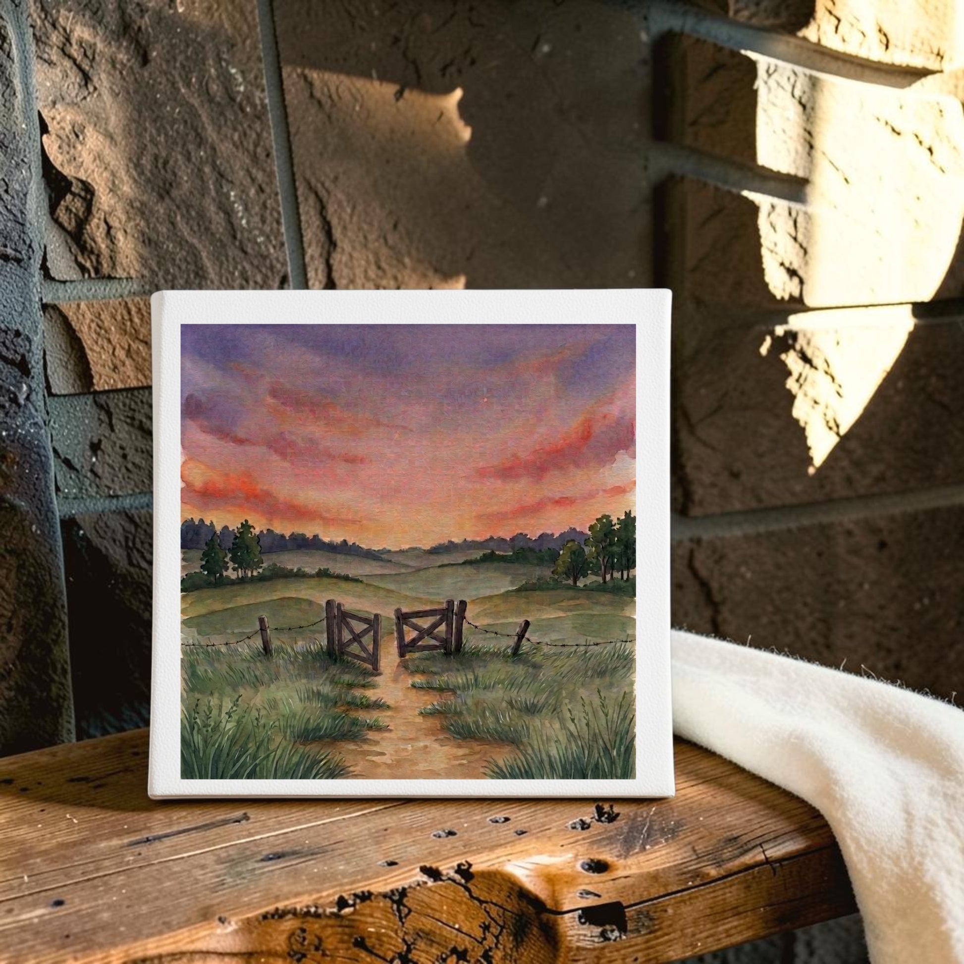 The “There Is More Sunset Landscape Canvas Art | 8x8” by Mirabilia Boutique depicts a serene country scene with a countryside path, wooden gate, and grassy fields illuminated by a vibrant sunset sky. Displayed on a wooden bench near a stone wall and a white cloth, this canvas artwork brings rustic charm to any space.