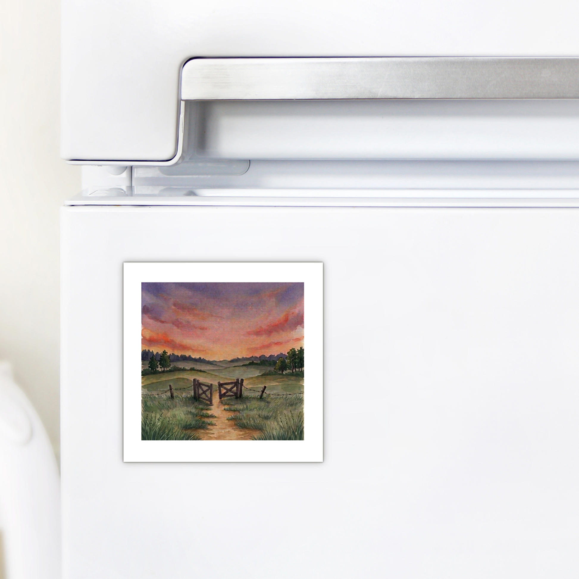 A white refrigerator with Mirabilia Boutique’s “There Is More Sunset Landscape Magnet,” showcasing inspirational art of a dirt path, grassy field, wooden fence, and a colorful sunset sky.