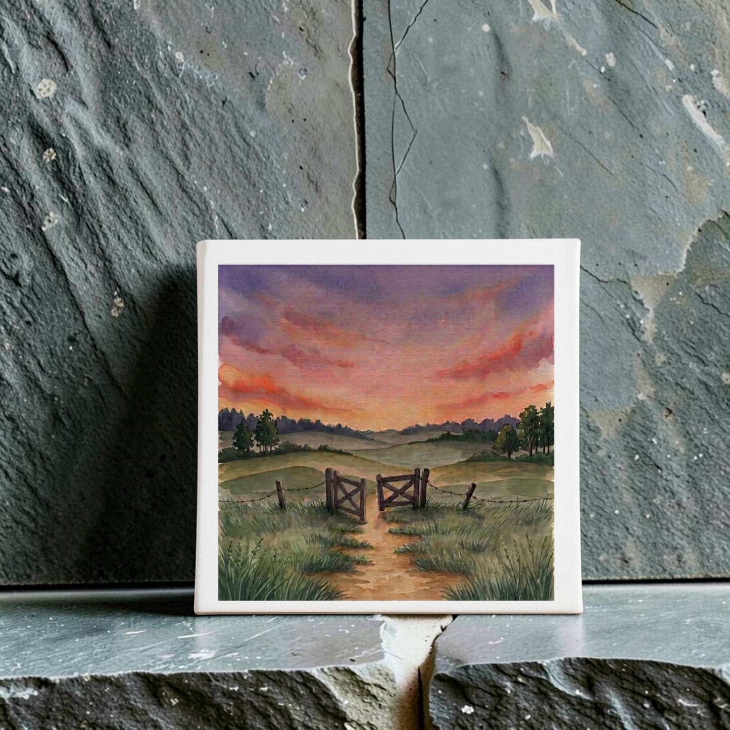 The “There Is More Sunset Landscape Canvas Art | 8x8” by Mirabilia Boutique features a striking country scene with a dirt path winding through an open wooden gate toward distant hills, surrounded by grassy fields and trees beneath a dramatic, colorful sunset sky. Displayed against a textured stone background, this artwork beautifully captures the tranquil beauty of the countryside at dusk.