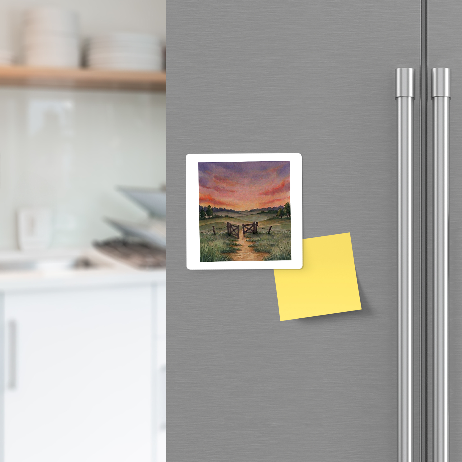 A refrigerator door with the Mirabilia Boutique “There Is More” Sunset Landscape Magnet and a blank yellow sticky note attached. Kitchen shelves and dishes are visible in the softly blurred background.