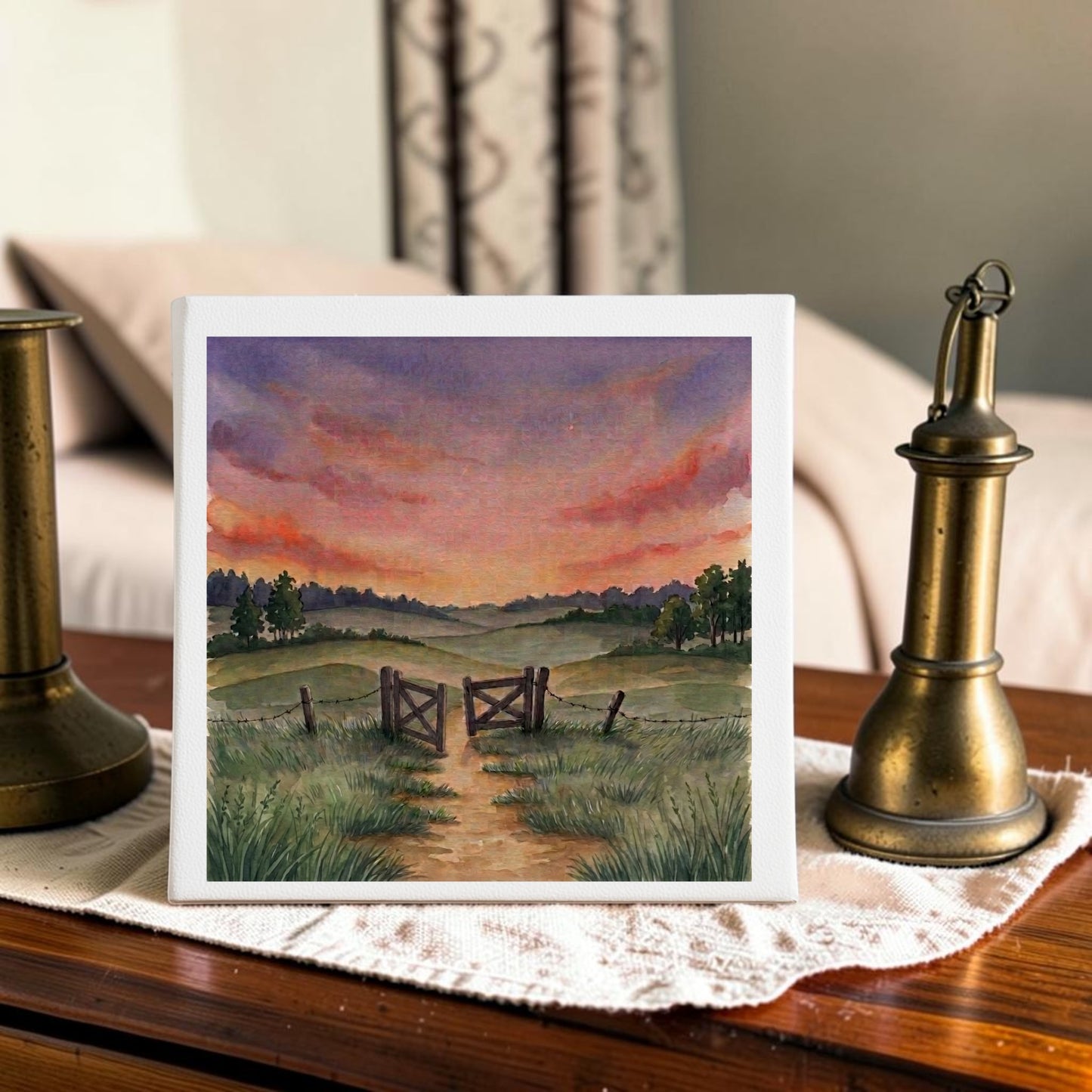 The “There Is More Sunset Landscape Canvas Art” (8x8) by Mirabilia Boutique, featuring a country path with a wooden gate and fence leading to grassy fields beneath a vibrant sunset sky, is displayed on a lace cloth between two brass candlesticks atop a wooden table.