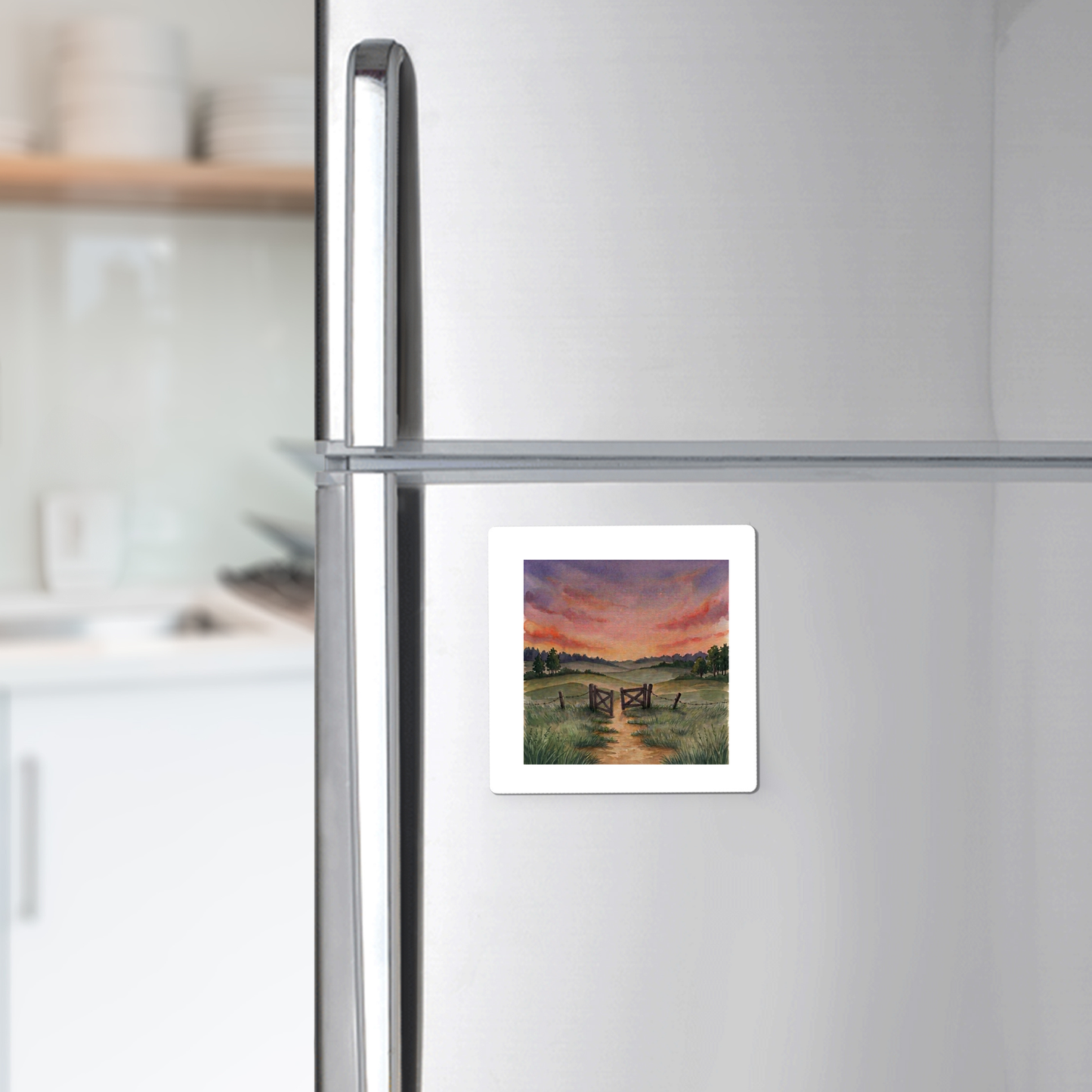 A “There Is More Sunset Landscape Magnet | Inspirational Art” by Mirabilia Boutique, featuring a rural sunset scene with a country path and wooden gate, is displayed on a stainless steel refrigerator in a modern kitchen.