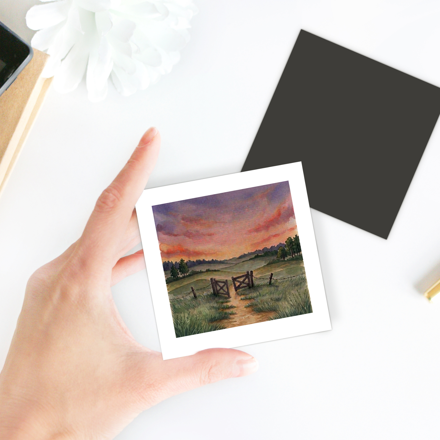 A hand holds a square “There Is More Sunset Landscape Magnet | Inspirational Art” by Mirabilia Boutique, featuring a rural landscape with a wooden gate, grassy path, and vibrant sunset sky; the charming country scene is beautifully captured on this magnet, while a black square card rests nearby on the white surface.