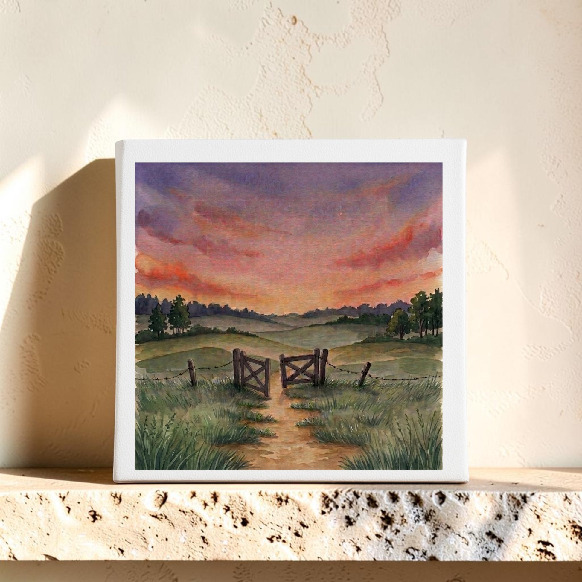 Display the “There Is More Sunset Landscape Canvas Art | 8x8” by Mirabilia Boutique—an artwork illustrating a countryside path winding toward a wooden gate amid grassy fields and trees beneath a vibrant sunset sky, perfectly propped on a textured stone shelf.