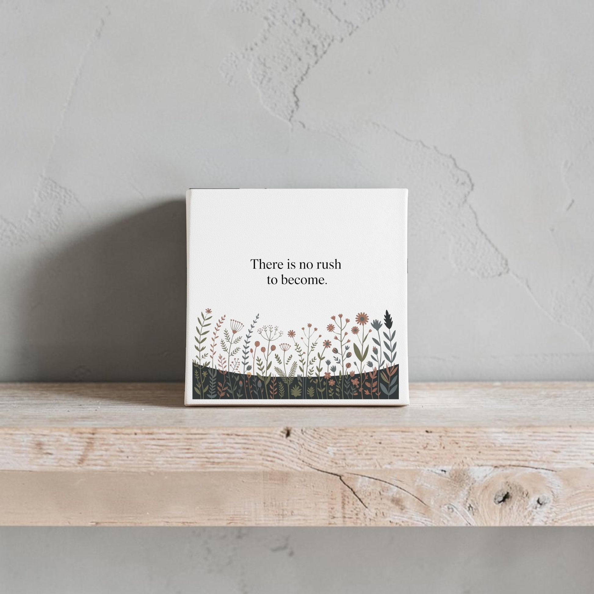 The Mirabilia Boutique "There Is No Rush to Become" Canvas Art features floral accents and adds motivational charm to your home décor, displayed here on a light wood shelf against a textured gray wall.
