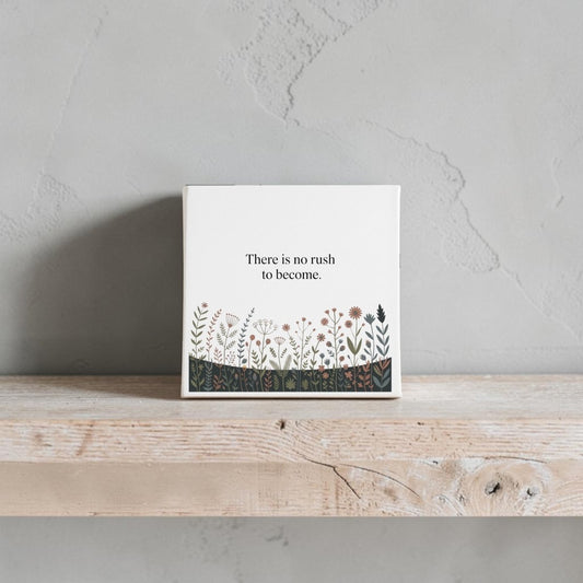 The Mirabilia Boutique "There Is No Rush to Become" Canvas Art features floral accents and adds motivational charm to your home décor, displayed here on a light wood shelf against a textured gray wall.