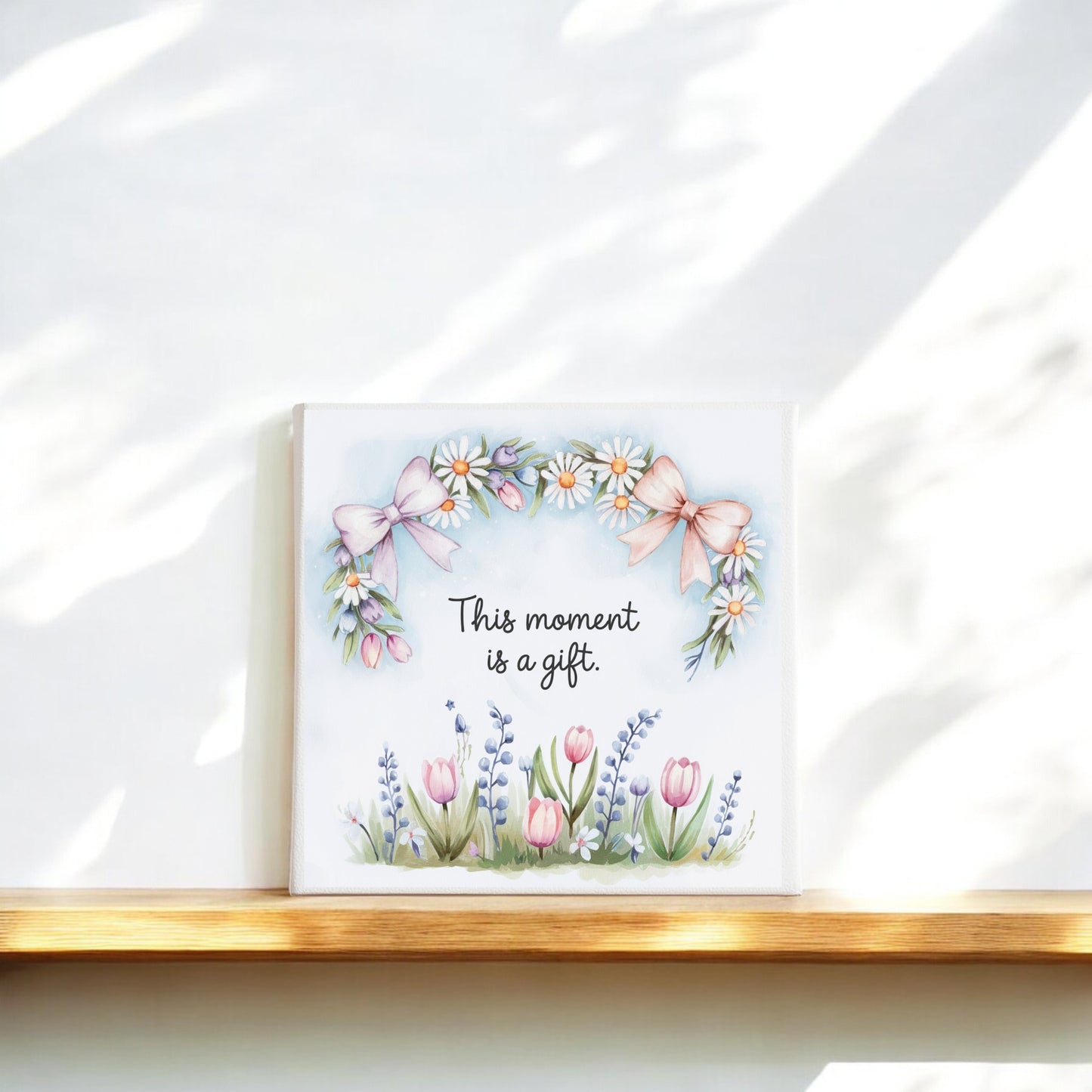 The Mirabilia Boutique "This Moment Is a Gift Canvas – 8x8 Spring Floral Art" features delicate florals and pink bows, making it perfect for mindful wall art or slow living decor on any softly lit wooden ledge.