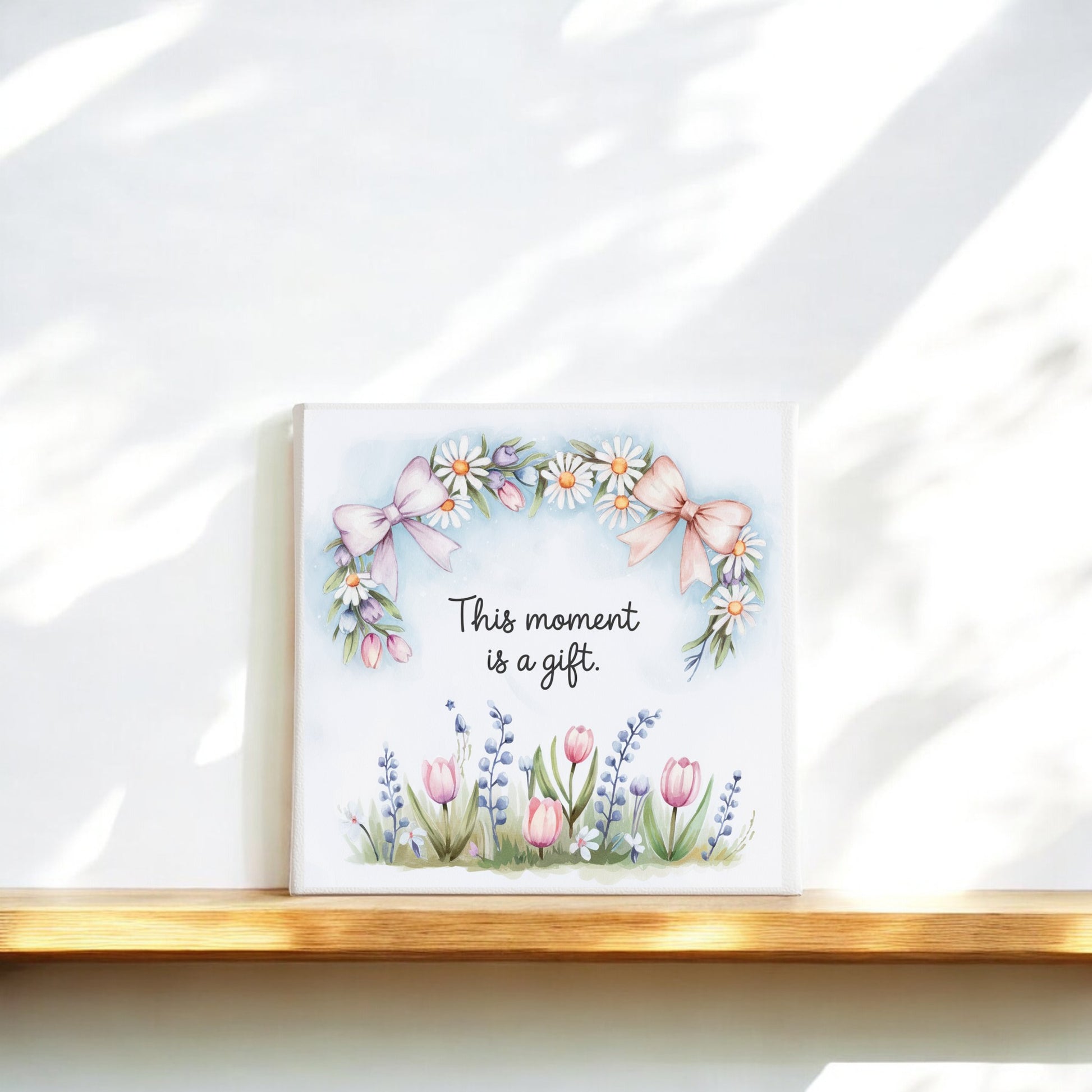 The Mirabilia Boutique "This Moment Is a Gift Canvas – 8x8 Spring Floral Art" features delicate florals and pink bows, making it perfect for mindful wall art or slow living decor on any softly lit wooden ledge.