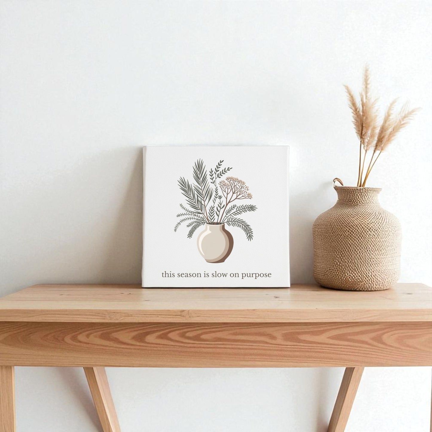 A minimalist wooden table displays a decorative vase and the "Slow on Purpose Canvas | Botanical Vase Quote Wall Art" by Mirabilia Boutique, embracing intentional living against a plain white wall.