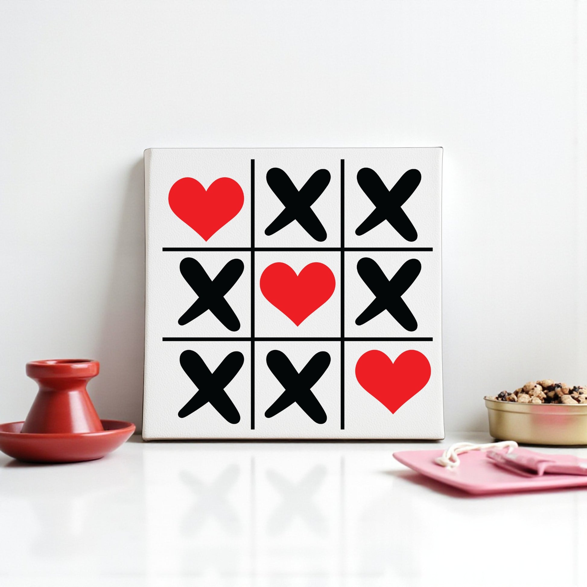 The Mirabilia Boutique Tic Tac Toe Love Sign is an 8x8 canvas wall art featuring red hearts and black Xs—three hearts and six Xs—displayed on a table with Valentine’s Day decor like a red candle holder, gold snack bowl, and pink plate with a fork.