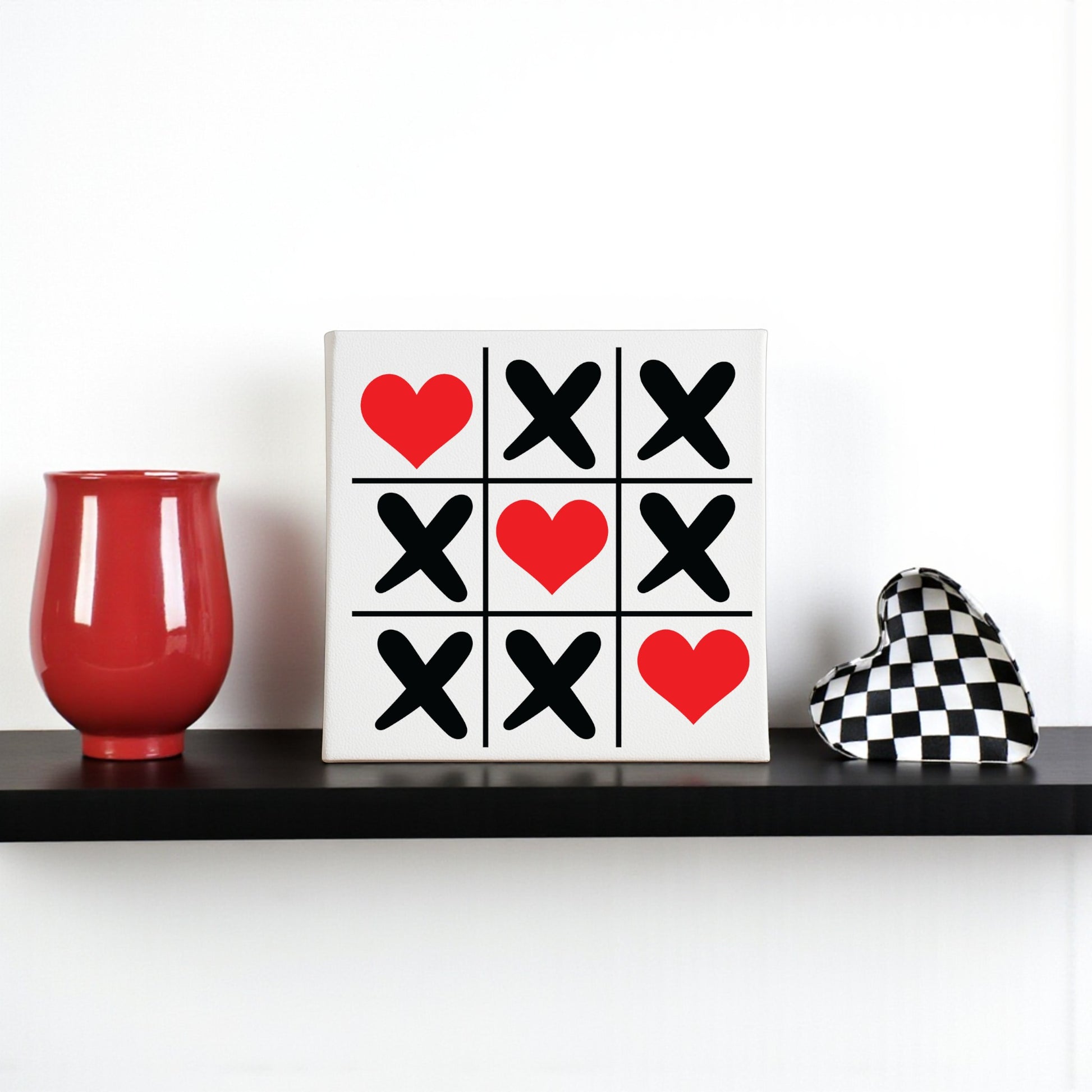 A shelf displays a red vase, a Mirabilia Boutique Tic Tac Toe Love Sign (8x8 Valentine Canvas Wall Art) with black X's and red hearts, and a black-and-white checkered heart—perfect for Valentine’s Day decor or heart wall art.