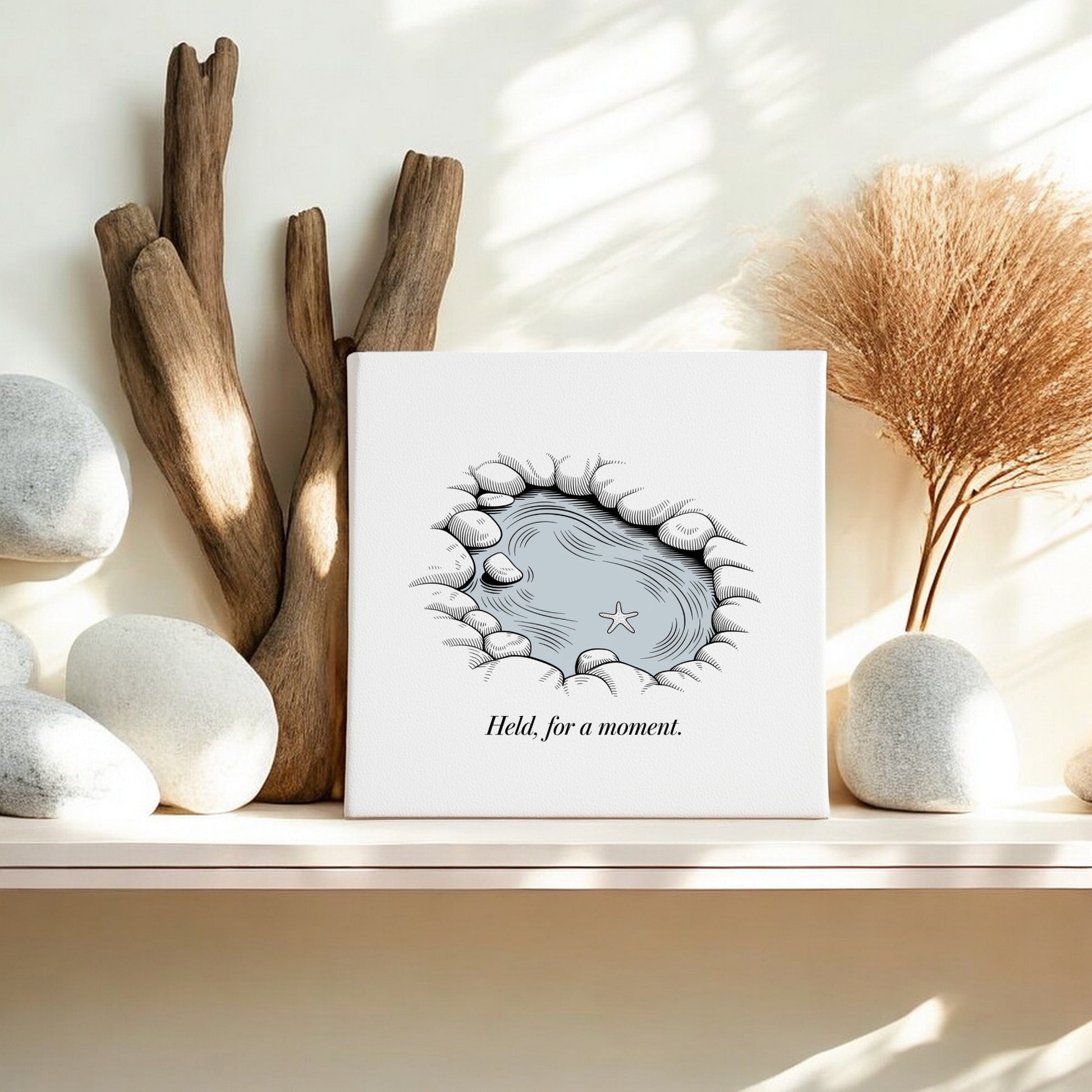 A shelf displays smooth white stones, driftwood, dried grass in a vase, and Mirabilia Boutique's "Held, For a Moment" – an 8x8 coastal canvas wall art featuring an illustrated tide pool.