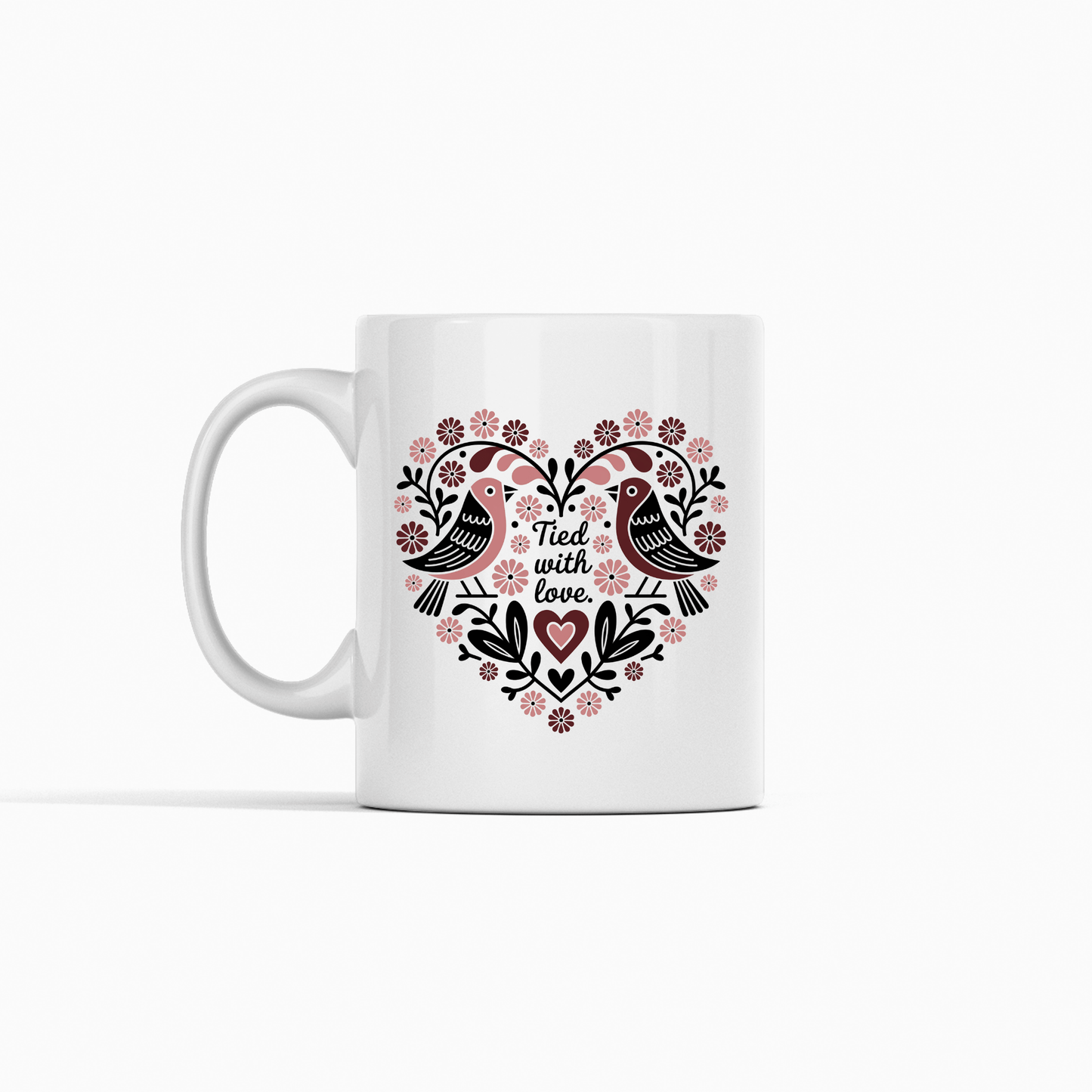 The Mirabilia Boutique "Tied With Love Mug – Folk Art Heart & Birds" is a white ceramic mug with black and pink accents, featuring love birds and a heart-shaped floral design with the phrase "Filled with love." A charming anniversary gift.