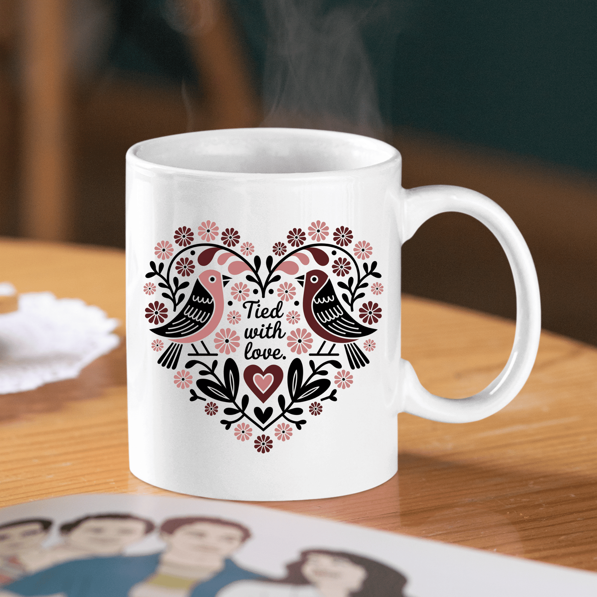 The Tied With Love Mug – Folk Art Heart & Birds by Mirabilia Boutique sits on a table, decorated with love birds, flowers, and leaves in a heart shape. Steam rises from the mug, making it a perfect anniversary gift.