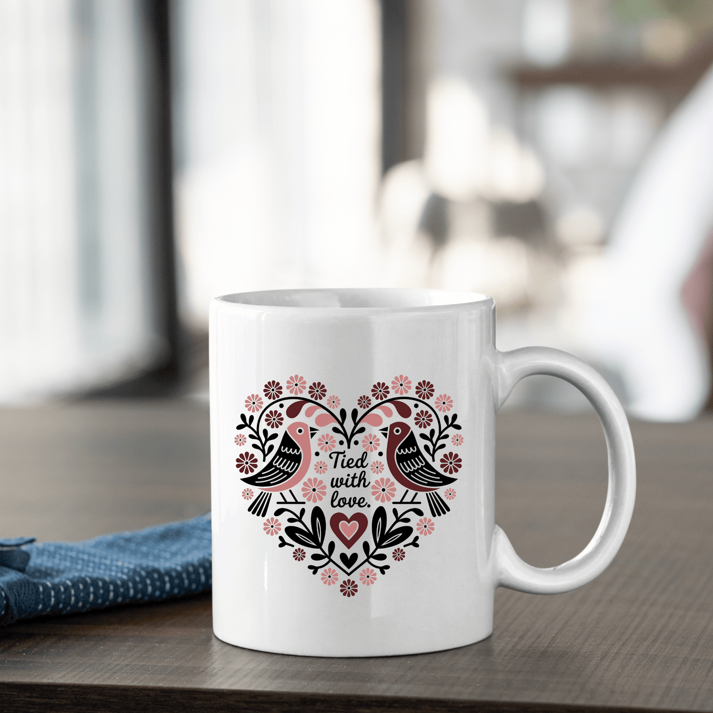 The Mirabilia Boutique "Tied With Love Mug – Folk Art Heart & Birds" features two love birds and floral heart on white ceramic, perfect as an anniversary gift. Shown on a wooden table with a folded blue cloth nearby.