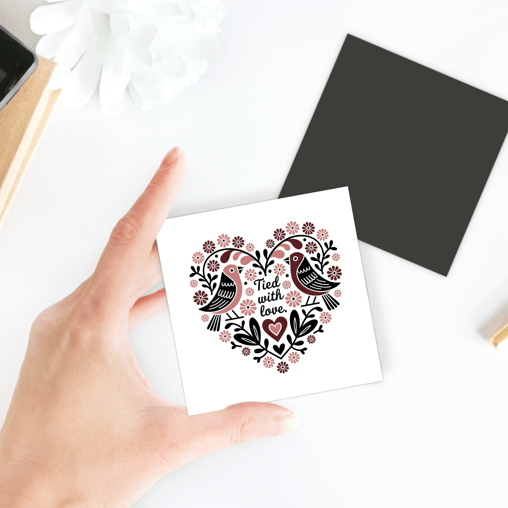 A hand displays the "Tied With Love Birds Magnet" by Mirabilia Boutique—a square magnet with two birds, flowers, and a heart reading "Tied with love." Shown against a white background with a black envelope and nearby flower—perfect as a wedding favor.