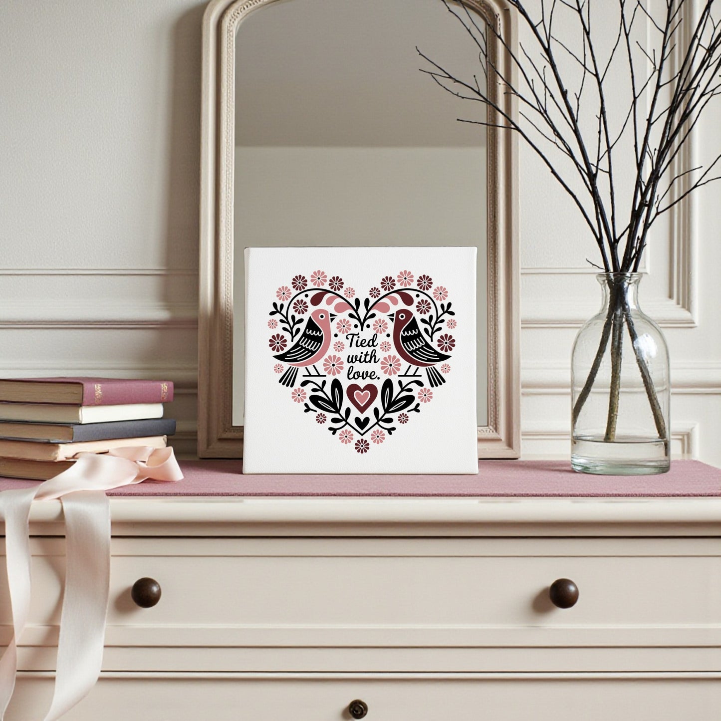 A dresser with a pink cloth holds stacked books, a ribbon, and a glass vase with bare branches. A Mirabilia Boutique Tied With Love Folk Art Valentine Canvas featuring birds and flowers decorates the mirror, adding charm.