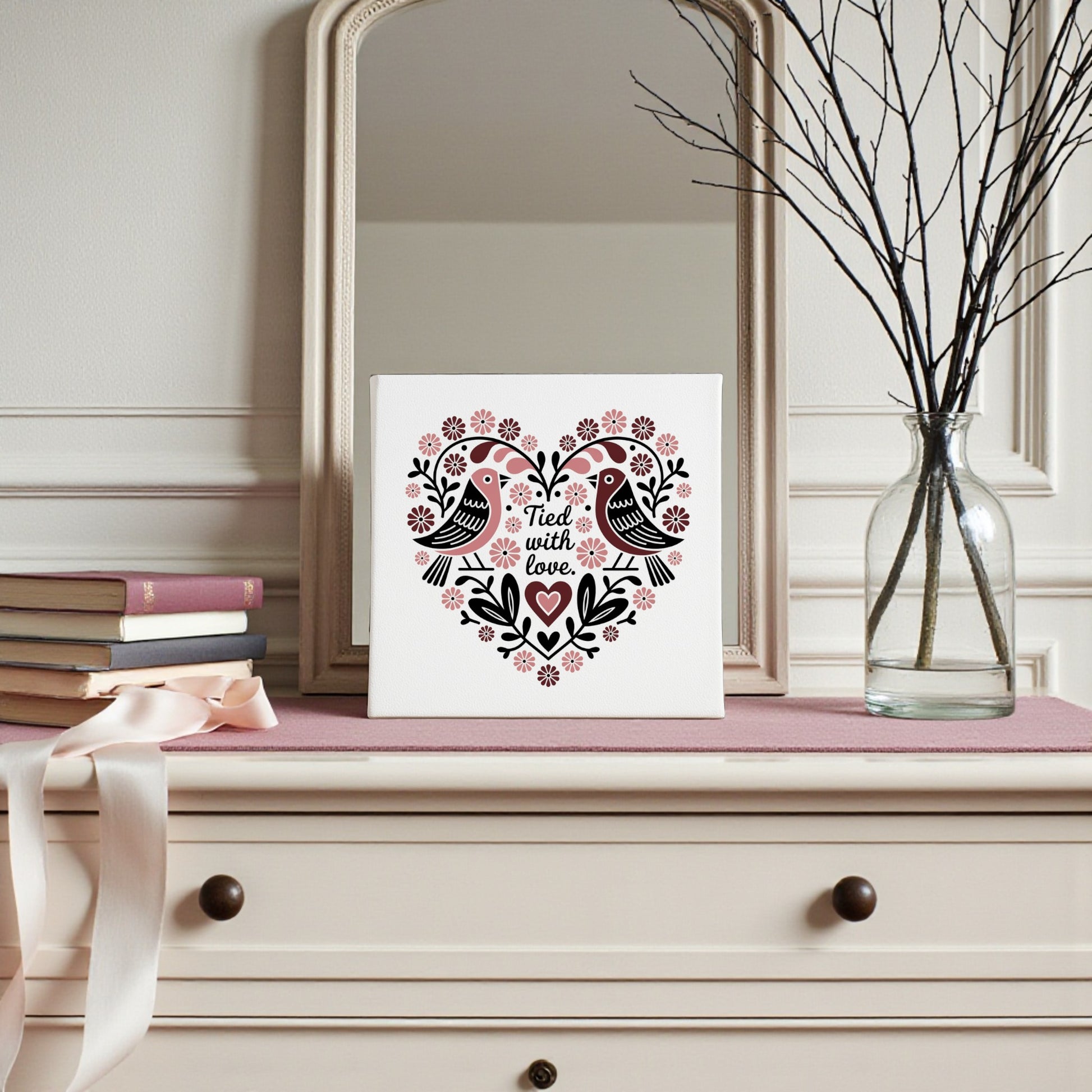 A dresser with a pink cloth holds stacked books, a ribbon, and a glass vase with bare branches. A Mirabilia Boutique Tied With Love Folk Art Valentine Canvas featuring birds and flowers decorates the mirror, adding charm.