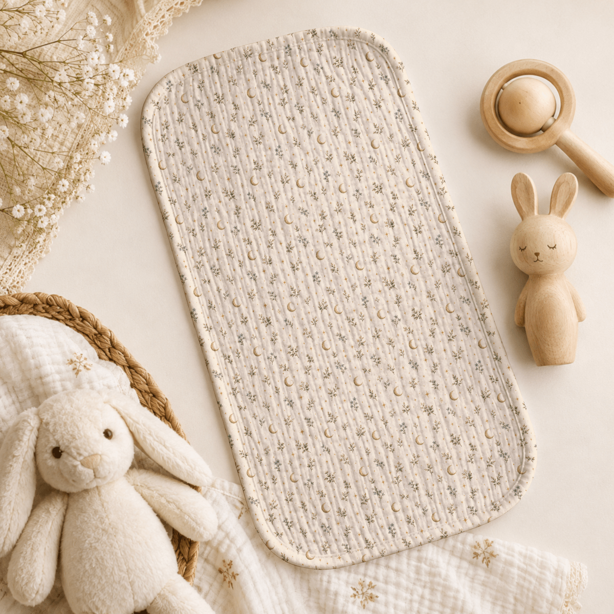 A quilted baby changing mat featuring a delicate floral motif—an essential botanical addition to your nursery—styled alongside a plush bunny, a wooden bunny toy, a wooden rattle, and a textured blanket on a soft neutral backdrop. Thoughtfully curated for the Moon & Moss Nursery by Mirabilia Boutique or beautifully paired with the Tiny Stars Burp Cloth.