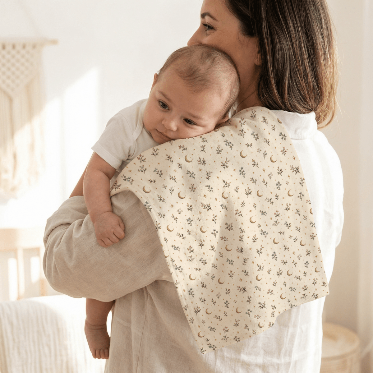 A mother in a light-colored top holds her baby on her shoulder. The baby, dressed in a white onesie, rests its head on the Tiny Stars Burp Cloth by Mirabilia Boutique—a botanical nursery essential from Moon & Moss Nursery—featuring a cream background adorned with small blue flowers, gold crescent moons, and stars.
