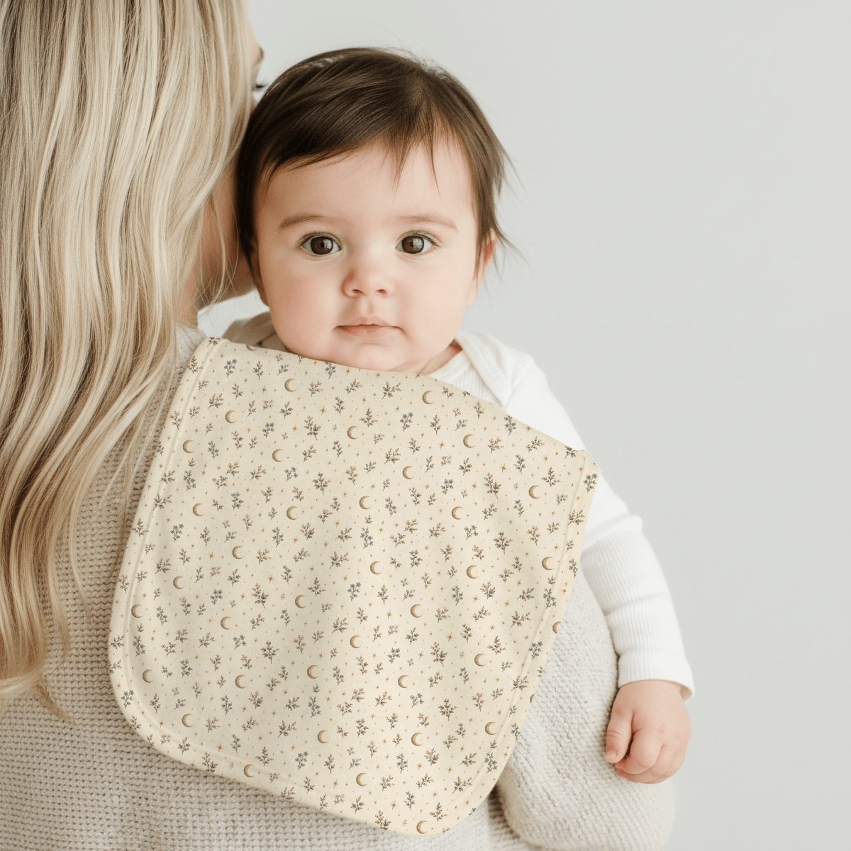 A baby with dark hair rests on an adult’s shoulder, dressed in a white outfit. Draped over the adult’s shoulder is the Tiny Stars Burp Cloth from Mirabilia Boutique’s Moon & Moss Nursery collection—a cream-colored essential featuring a charming star and moon pattern.