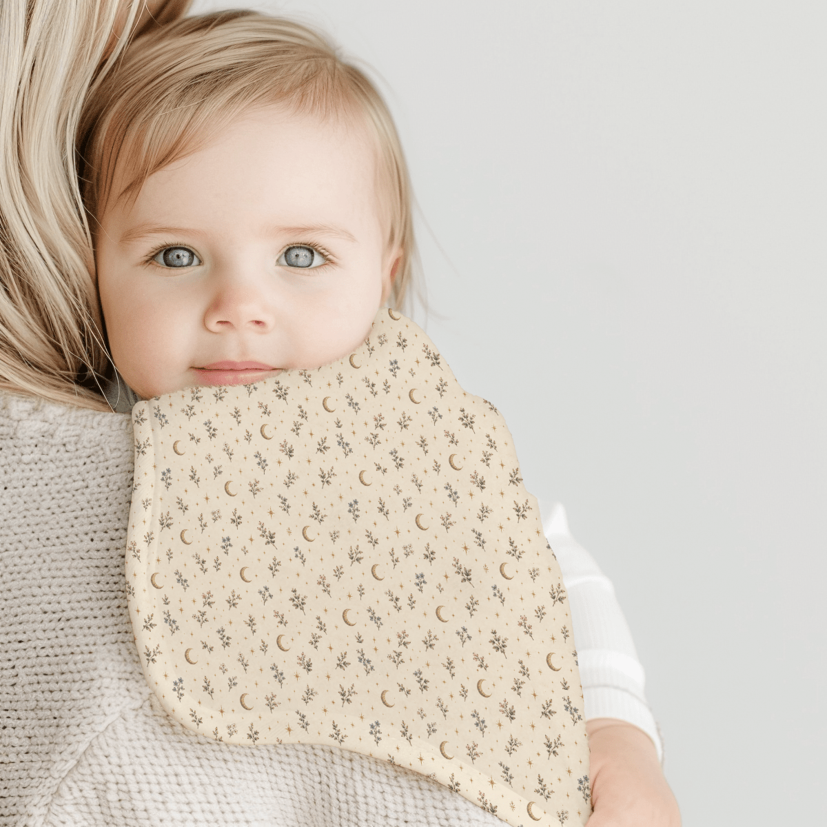 A baby with blue eyes rests on an adult’s shoulder, cuddling a Tiny Stars Burp Cloth from Mirabilia Boutique. The beige blanket, adorned with a moon and star pattern, enhances the soft and gentle atmosphere of the Moon & Moss Nursery collection. The serene scene is set against a light, neutral background.