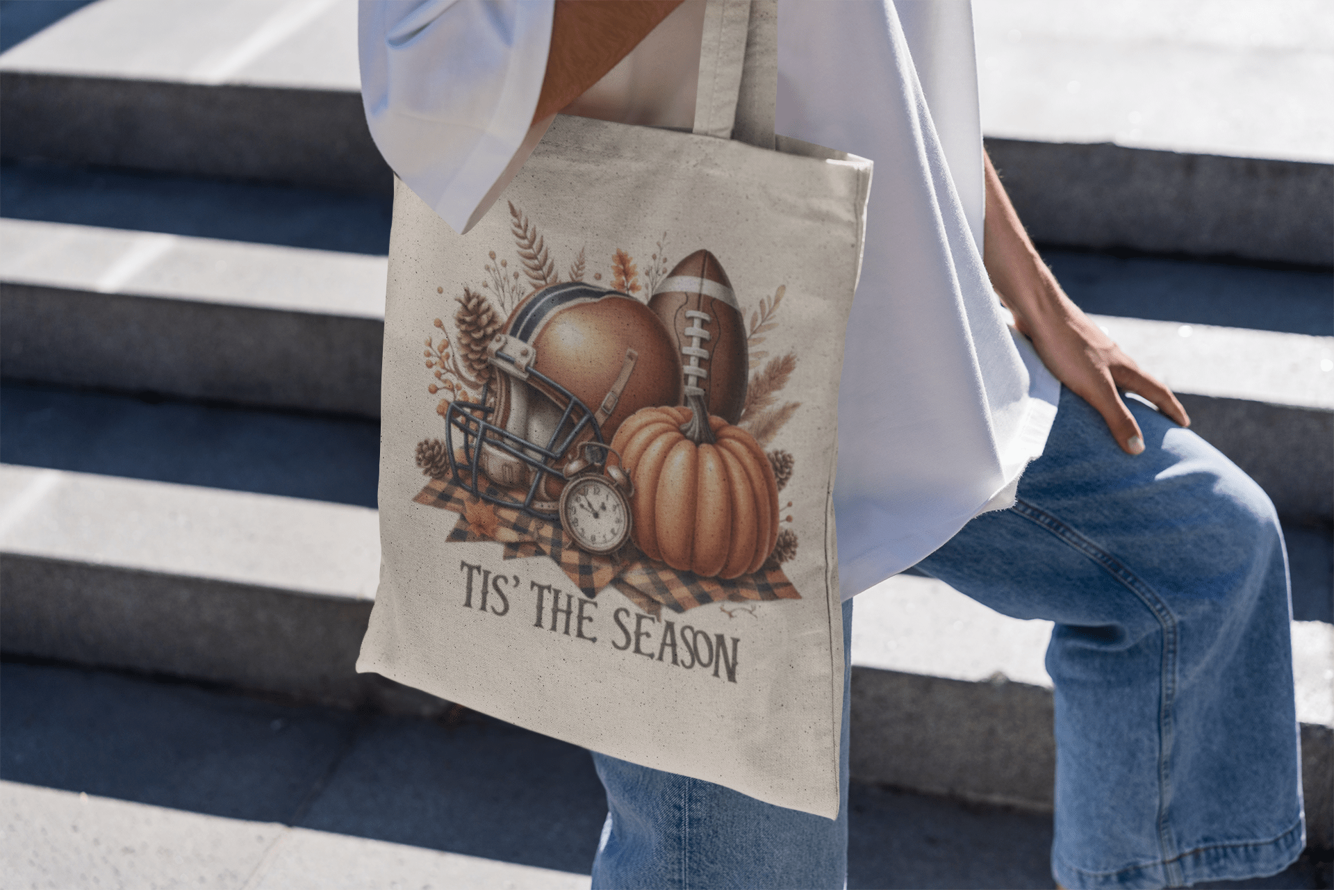 A person sits on outdoor steps holding the Mirabilia Boutique "Tis’ the Season Tote Bag," featuring a football helmet, pumpkin, leaves, plaid cloth, and the text "Tis' The Season.