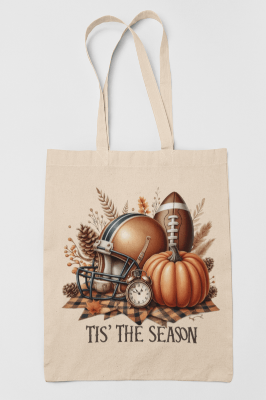The Mirabilia Boutique "Tis’ the Season Tote Bag" is a beige canvas market bag with autumn and football-themed illustrations, including a helmet, pumpkin, leaves, and plaid cloth, plus "TIS' THE SEASON" printed below.