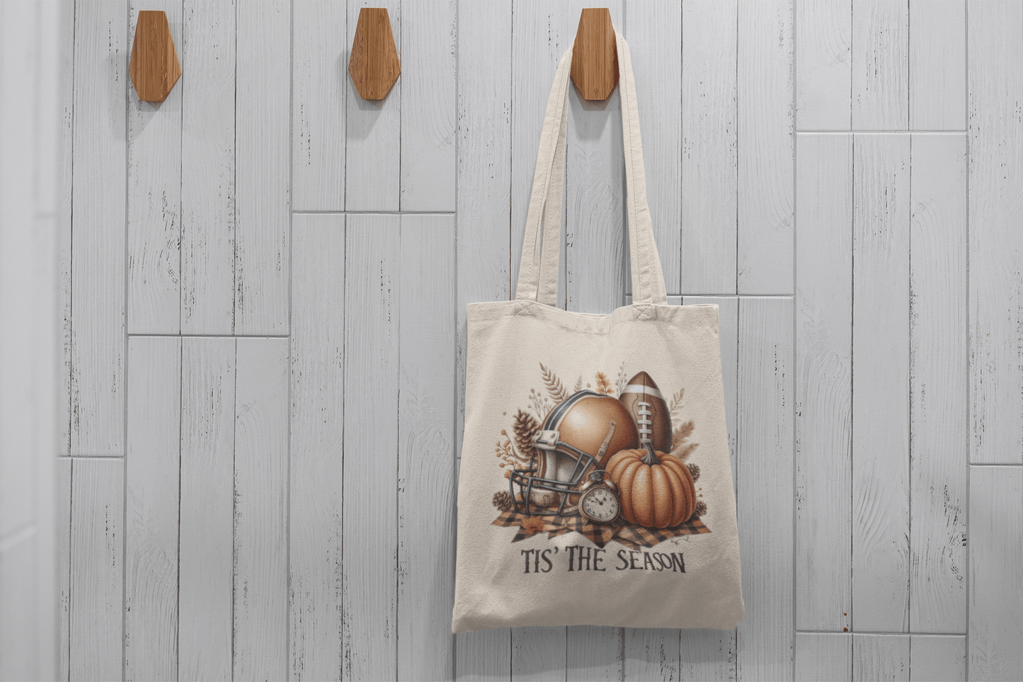 The Tis’ the Season Tote Bag by Mirabilia Boutique features football, pumpkin, and autumn motifs on beige fabric. Hanging on a wooden hook with a white wood panel background, it’s the perfect fall accessory for football fans.