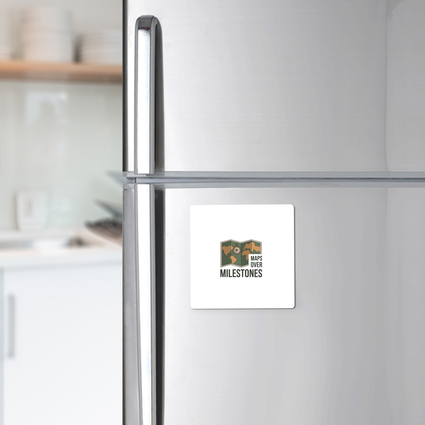 The Maps Over Milestones Travel Magnet by Mirabilia Boutique, featuring a logo with a map and the phrase "MAPS OVER MILESTONES," adds adventure-inspired style to any stainless steel fridge. Blurred kitchen shelves and dishes appear in the background.