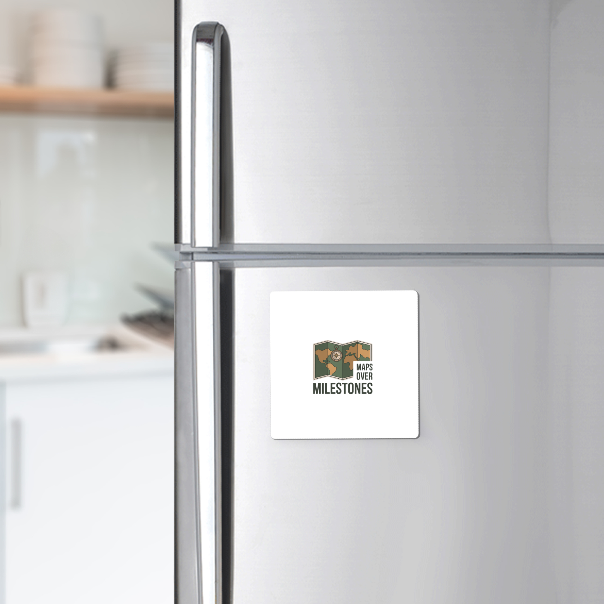 The Maps Over Milestones Travel Magnet by Mirabilia Boutique, featuring a logo with a map and the phrase "MAPS OVER MILESTONES," adds adventure-inspired style to any stainless steel fridge. Blurred kitchen shelves and dishes appear in the background.