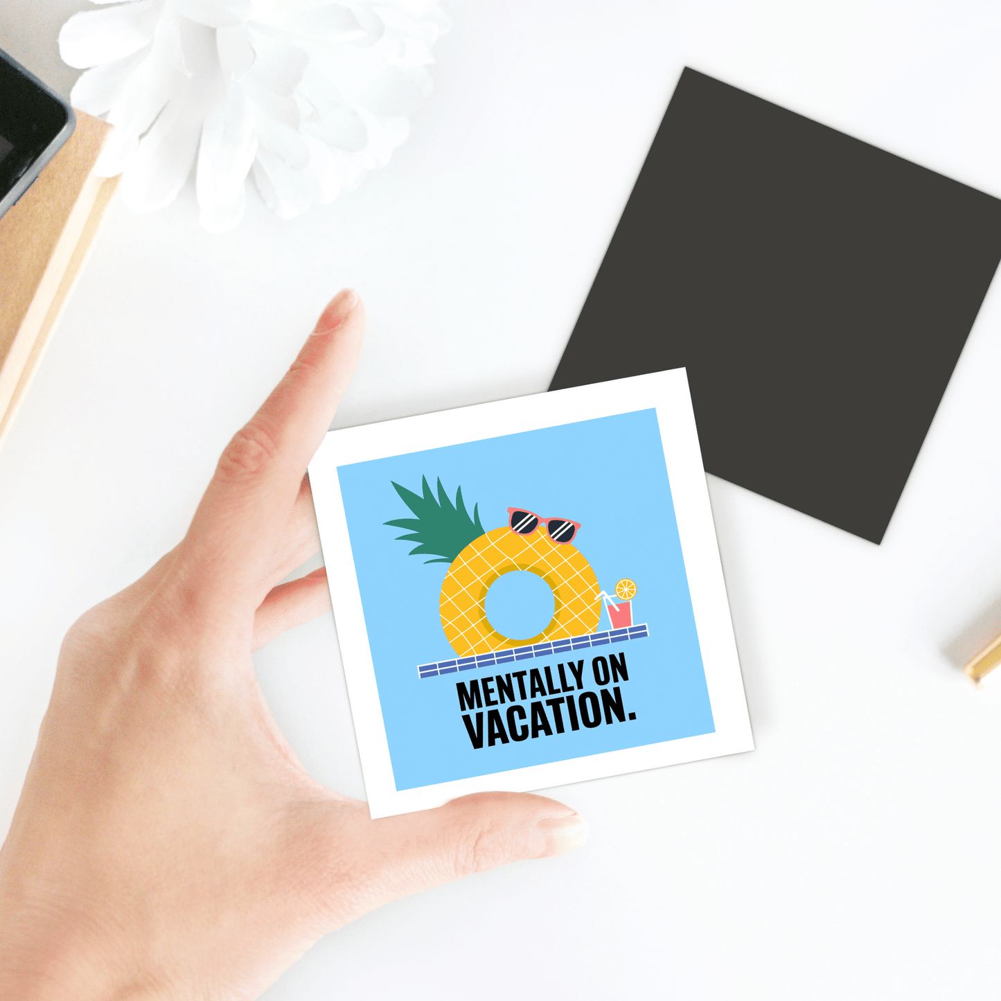A hand holds the Mirabilia Boutique "Mentally on Vacation Magnet – Tropical Summer Vibes," showing a pineapple with sunglasses and a drink. The magnet's card sits on a white surface beside a plant, black square, and other nearby items.