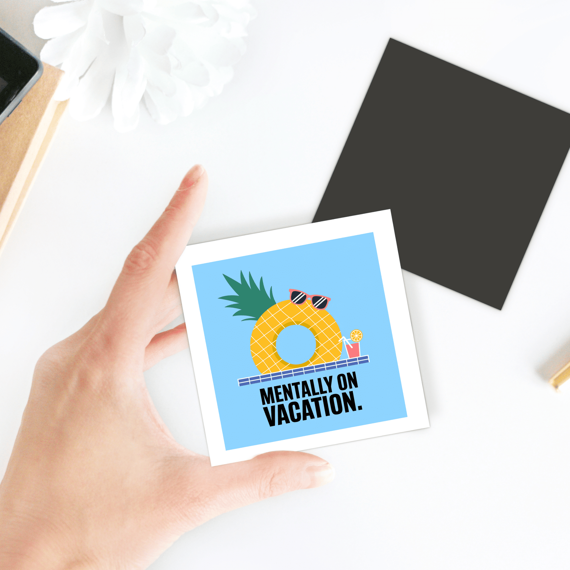 A hand holds the Mirabilia Boutique "Mentally on Vacation Magnet – Tropical Summer Vibes," showing a pineapple with sunglasses and a drink. The magnet's card sits on a white surface beside a plant, black square, and other nearby items.
