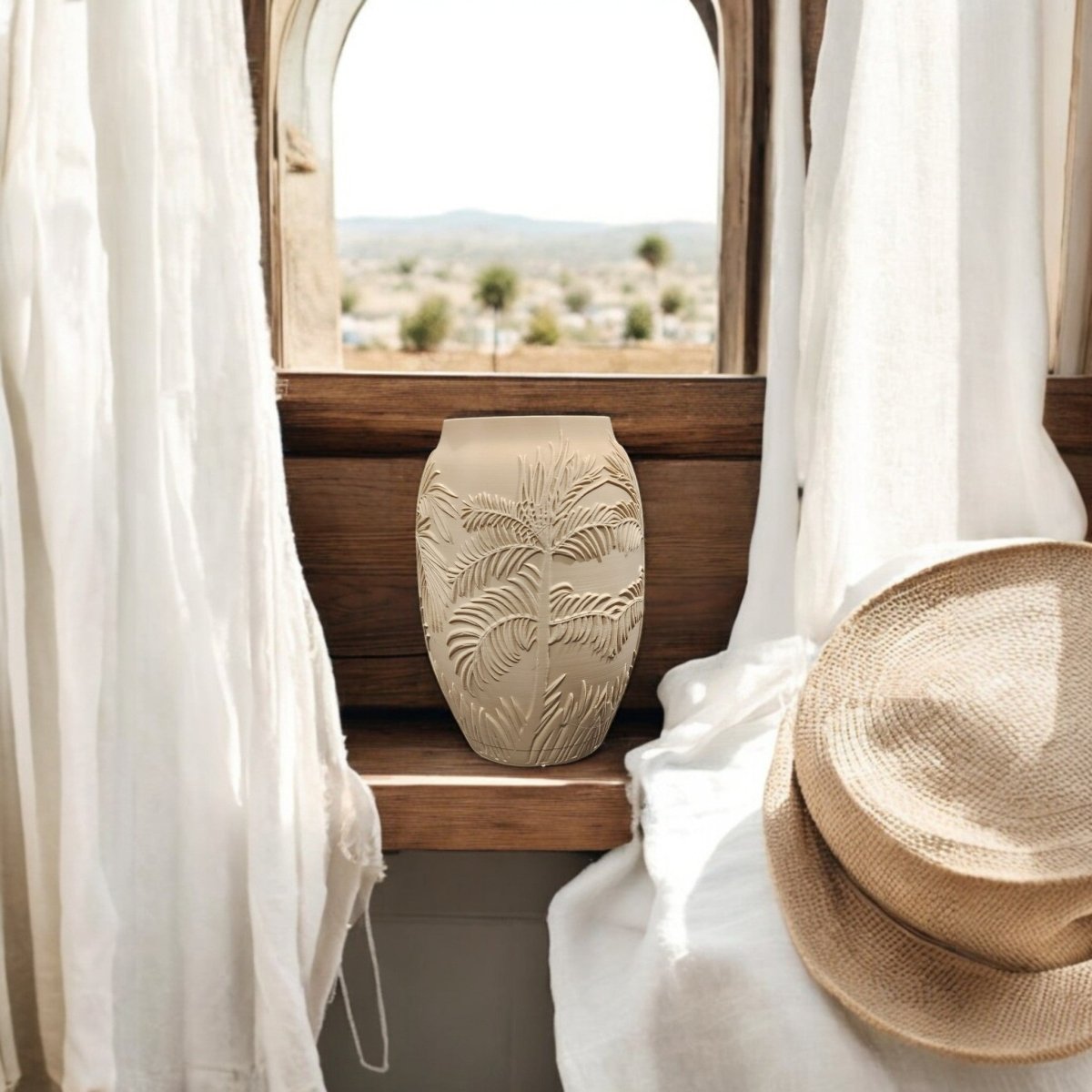 The Mirabilia Boutique Sand Palm Desert Vase for Summer Decor, featuring a palm leaf pattern, sits on a wooden windowsill between white curtains, next to a straw hat and an arched window overlooking trees and hills outside.