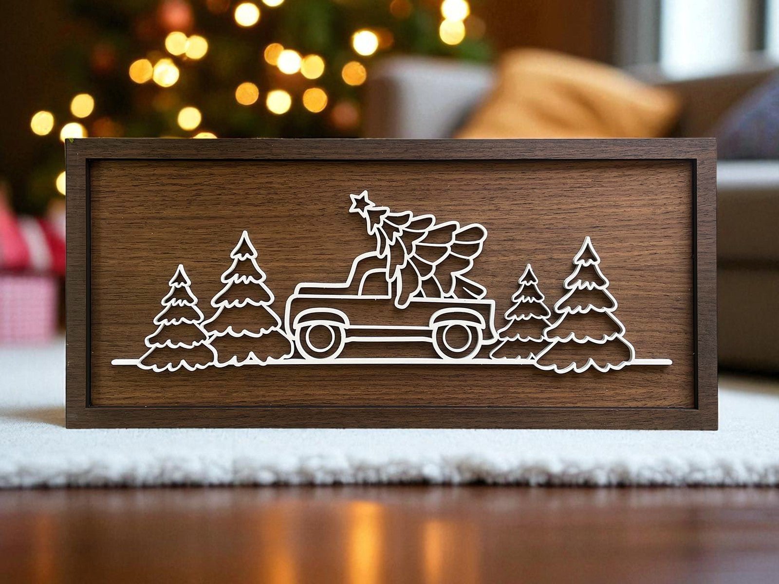 The Truck & Tree Laser Cut Christmas Sign by Mirabilia Boutique (18x8) features a white truck with a tree, surrounded by pines, set against blurred holiday lights for rustic seasonal decor.