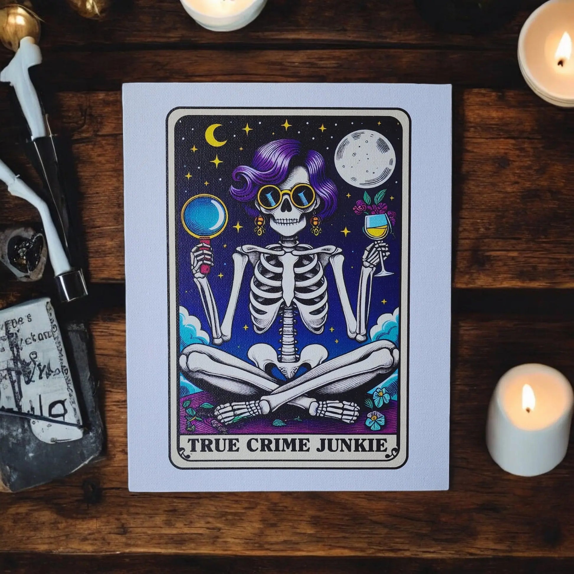 A framed 8x10 canvas sign from Mirabilia Boutique, titled "True Crime Junkie," displays a purple-haired skeleton with sunglasses, examining a magnifying glass and holding a wine glass. Surrounded by stars, a crescent moon, clouds, and flowers, the canvas sits on a wooden table with lit candles placed around it.