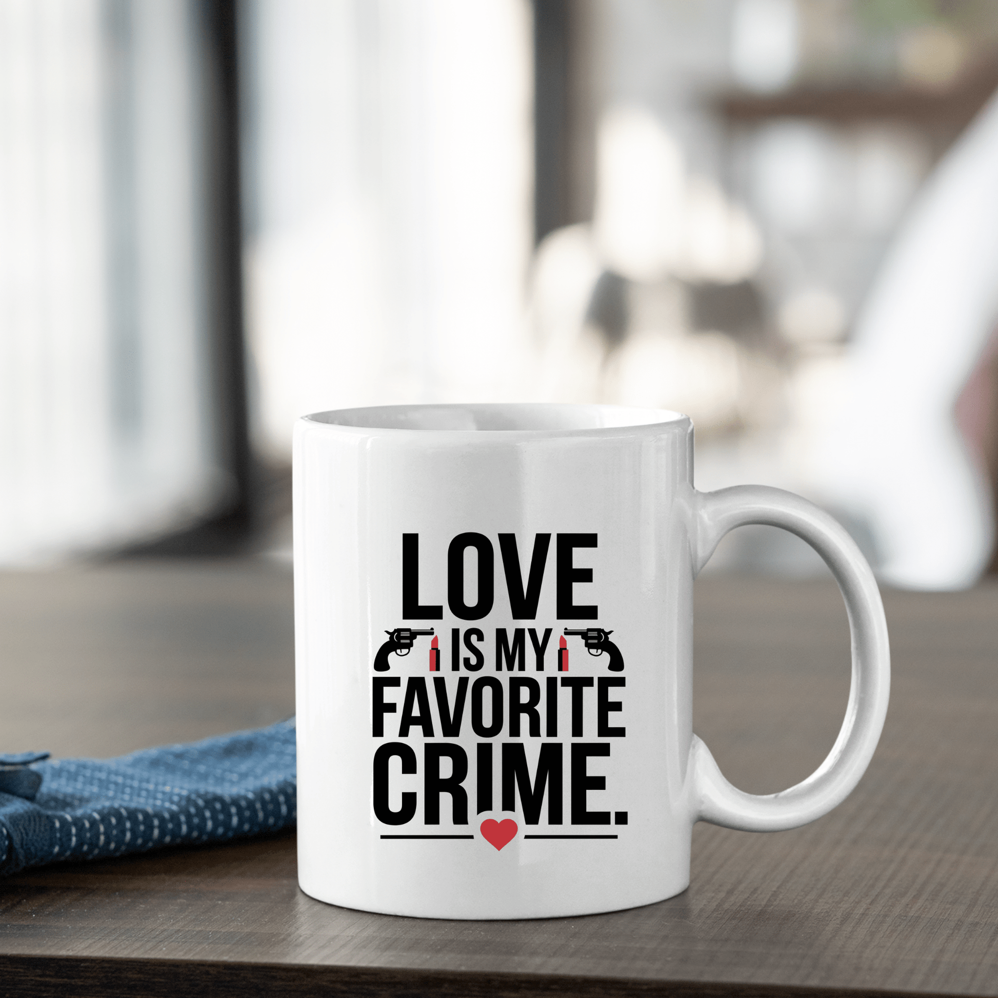 The Love Is My Favorite Crime Mug by Mirabilia Boutique sits on a wooden table with bold lettering, gun and handcuff icons, and a red heart—perfect true crime Valentine gift for podcast fans. A blue napkin is folded beside it.