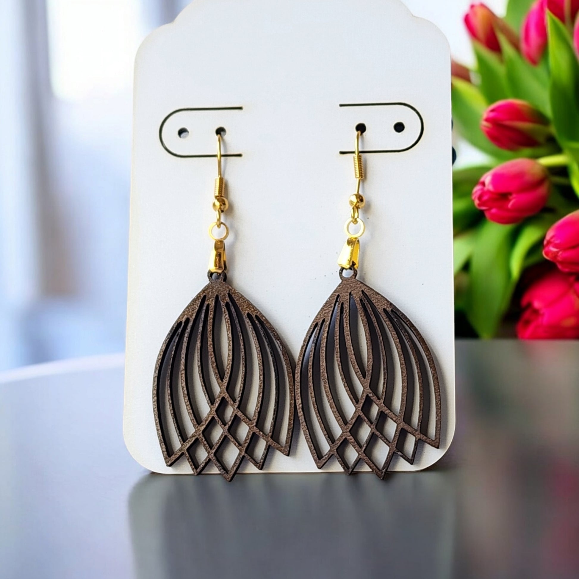 Mirabilia Boutique's Graceful Laser Cut Tulip Earrings, showcasing a delicate tulip design in layered, openwork metal, hang elegantly on a display card with gold hooks. In the background, softly focused red tulips with green leaves enhance their allure.