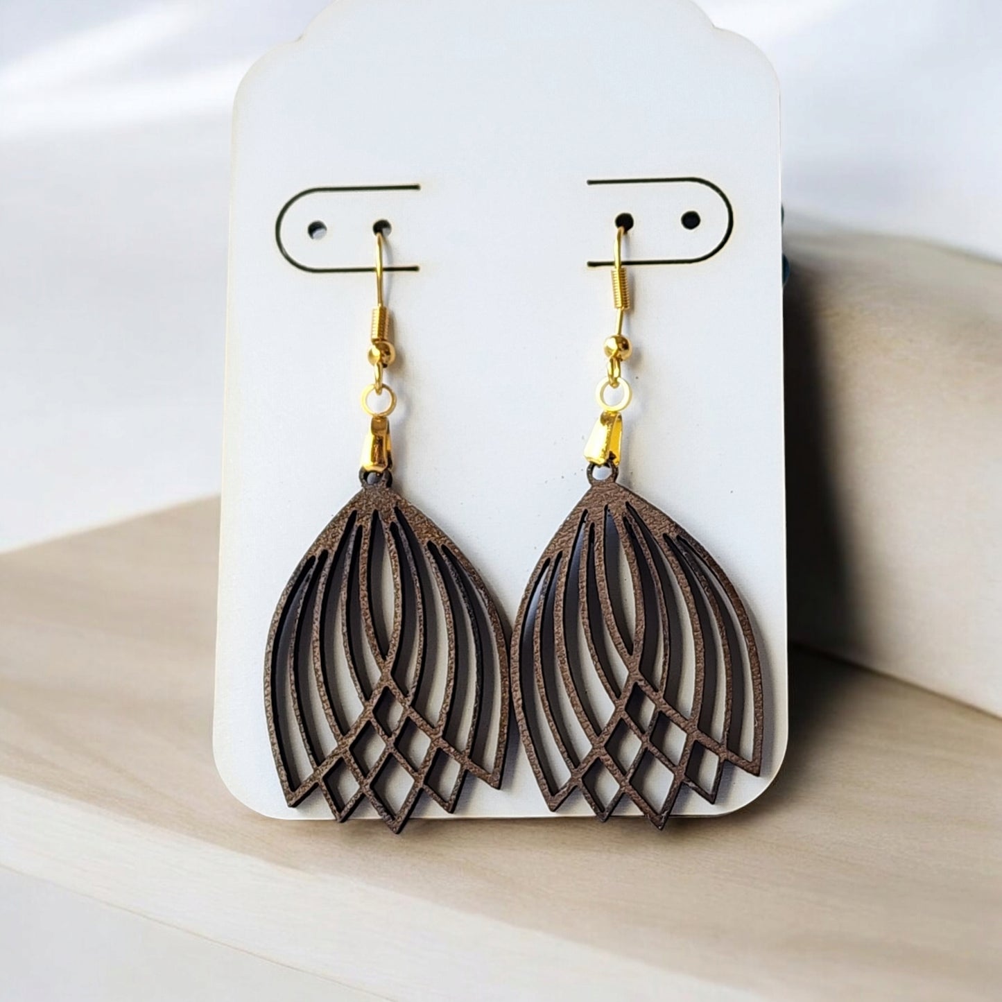 The Graceful Laser Cut Tulip Earrings from Mirabilia Boutique hang from a white display card. These handcrafted wooden earrings feature gold hooks and highlight the natural wood texture on a light wooden surface, adding an elegant touch with their tulip design.