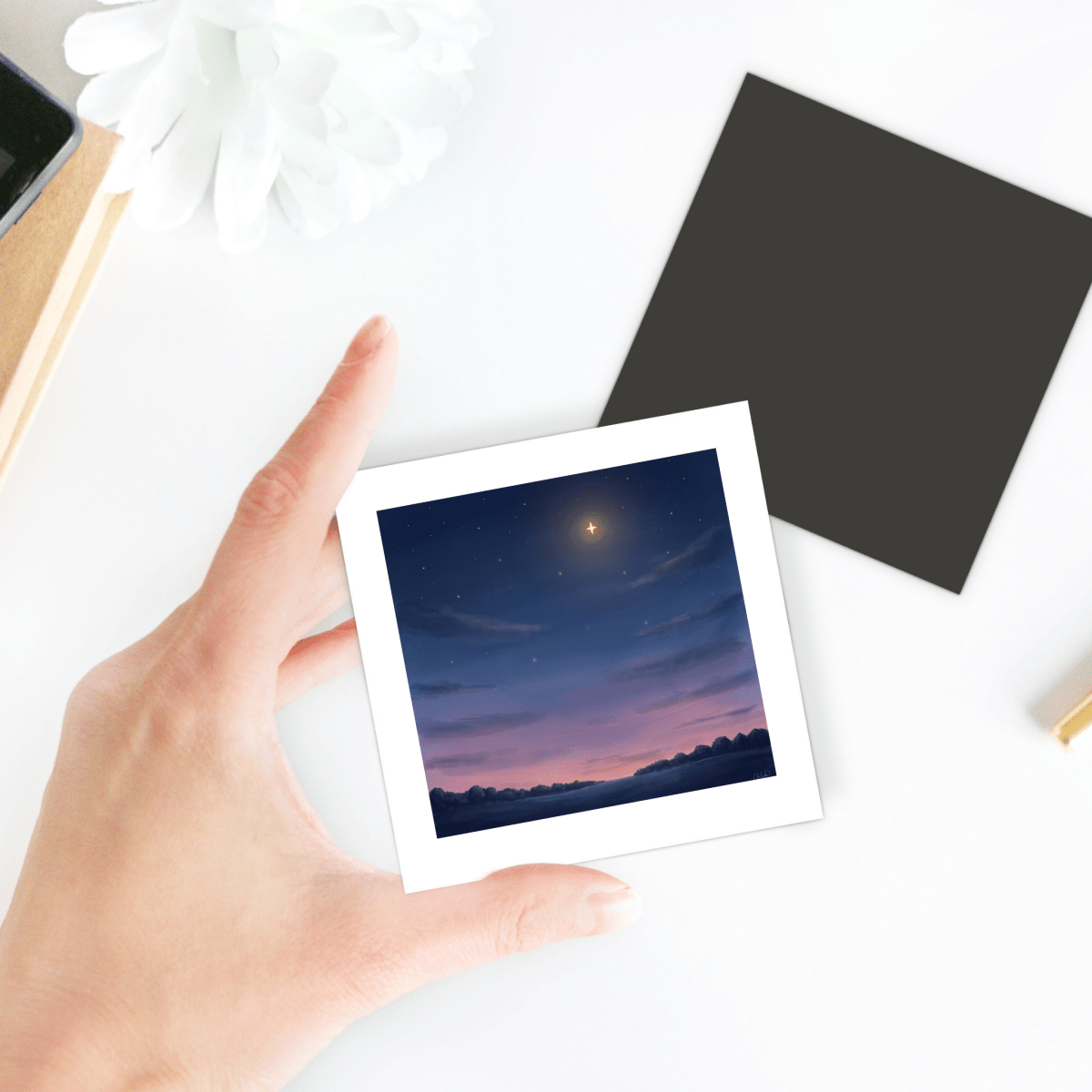 A hand holds a photo of a sunset sky with stars and a bright planet above silhouetted trees, featured on the Look Up Celestial Magnet by Mirabilia Boutique; items and a flower rest on a white table in the background, enhancing the charm of this decorative magnet.